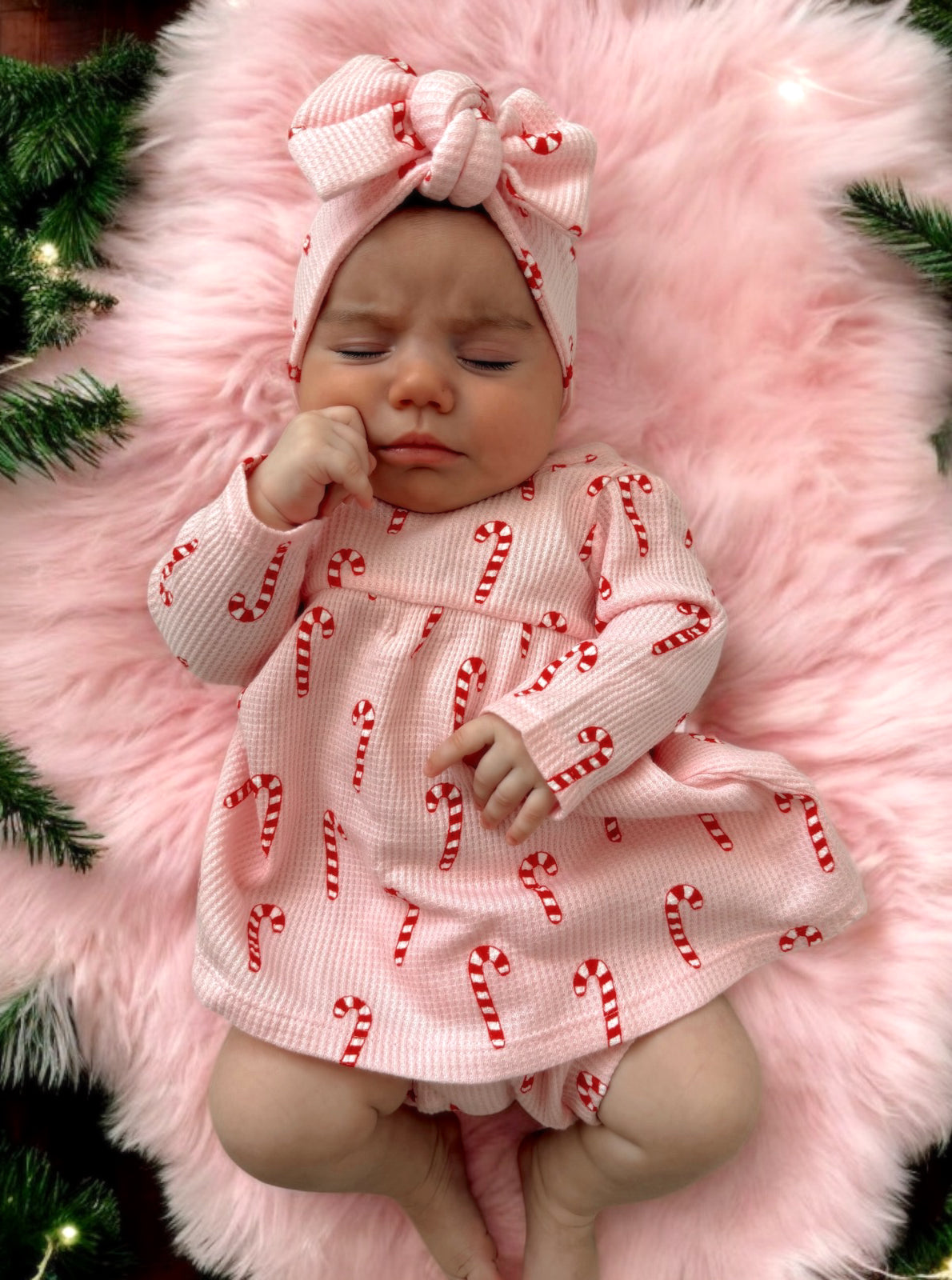 Baby girl in candy cane dress and headband, lying on pink fur with holiday greenery in the background.