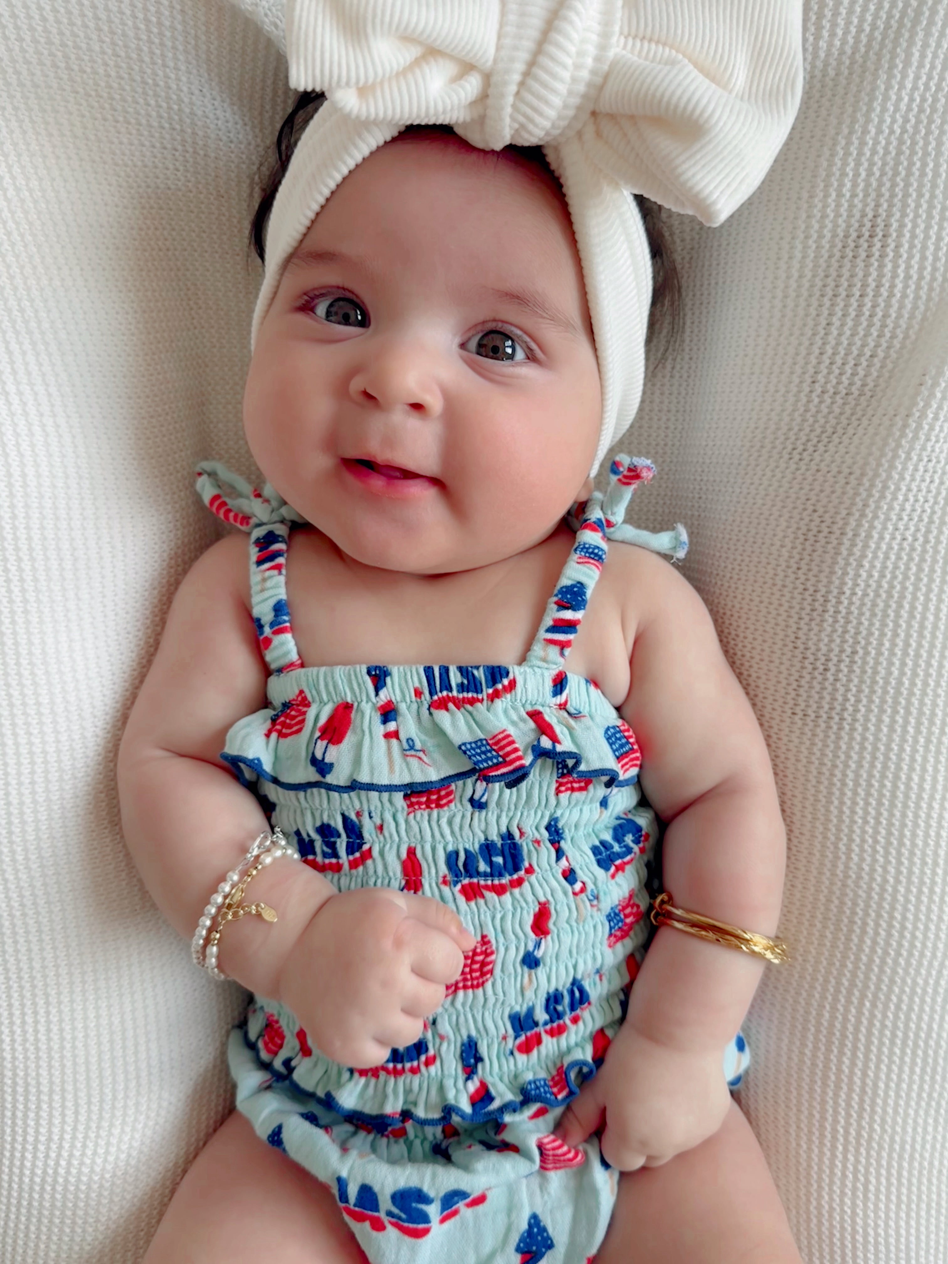 Smiling baby in a patterned romper with a large bow headband and bracelets, seated on a textured blanket.