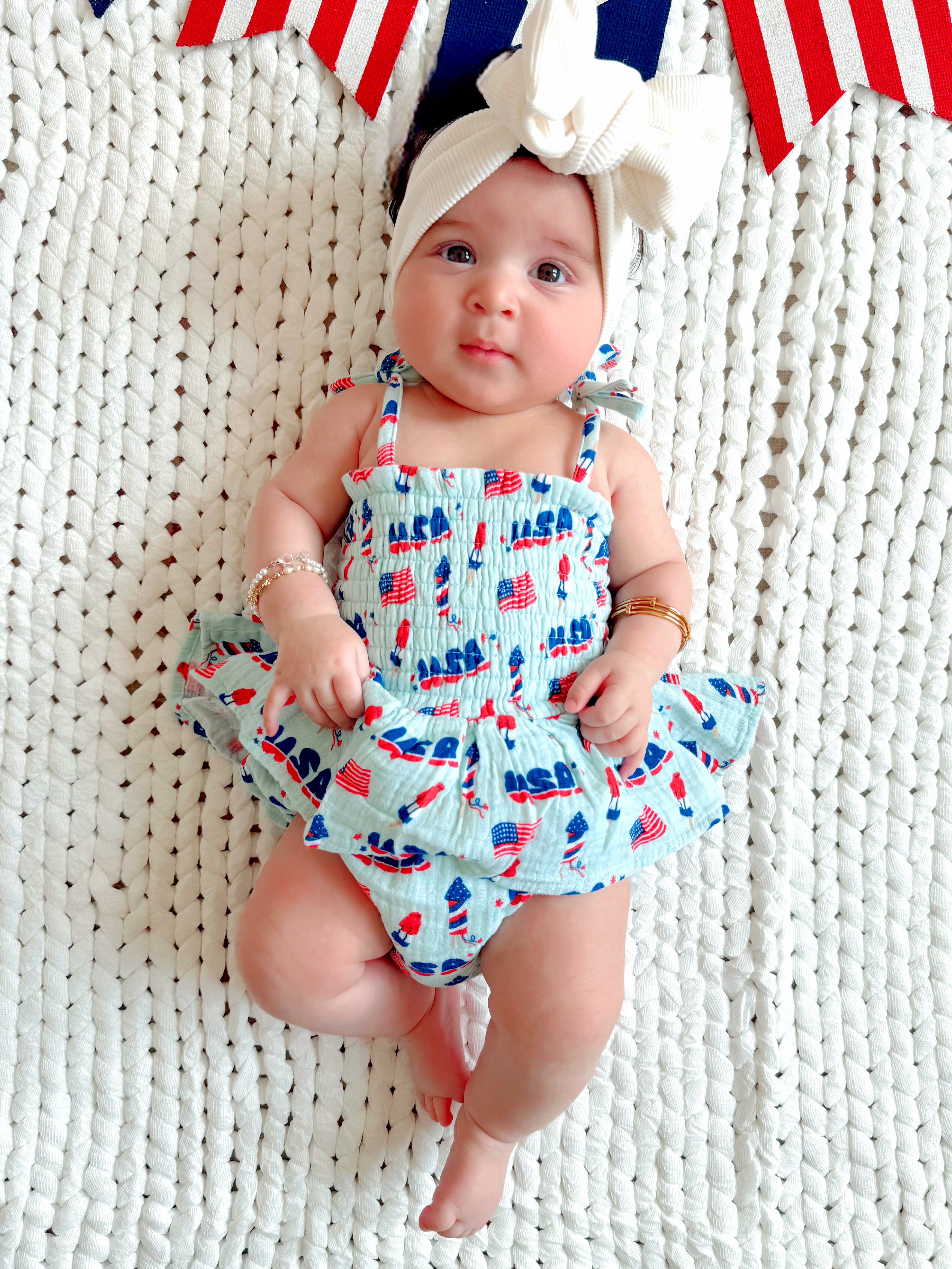 Baby girl in a patriotic outfit with a bow, lying on a textured blanket with red, white, and blue decorations.