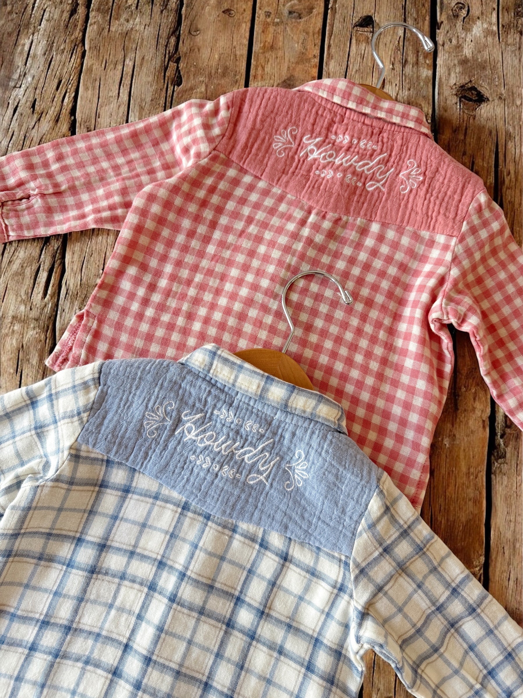 Two plaid shirts hanging on hooks, one pink and one blue, both featuring "Howdy" embroidery on the back.