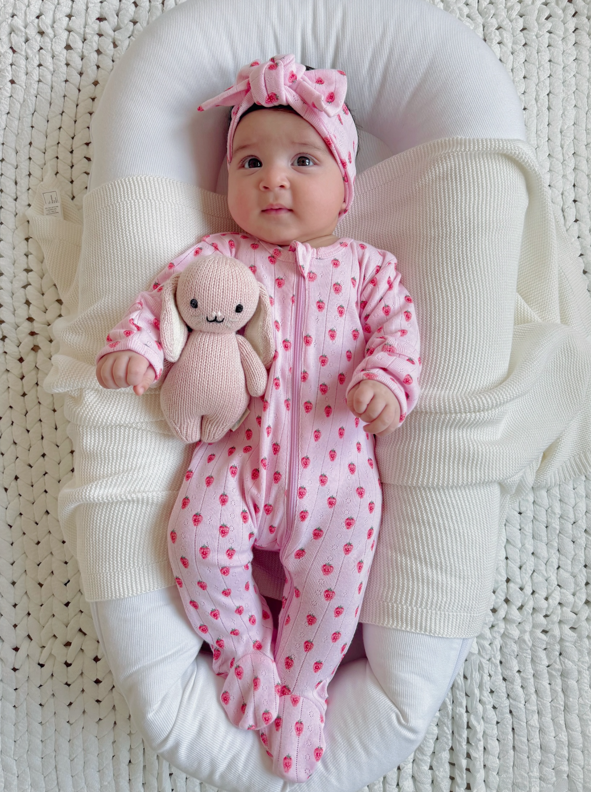 Baby girl in pink strawberry outfit holding a soft bunny, resting on a cozy blanket in a modern bassinet.