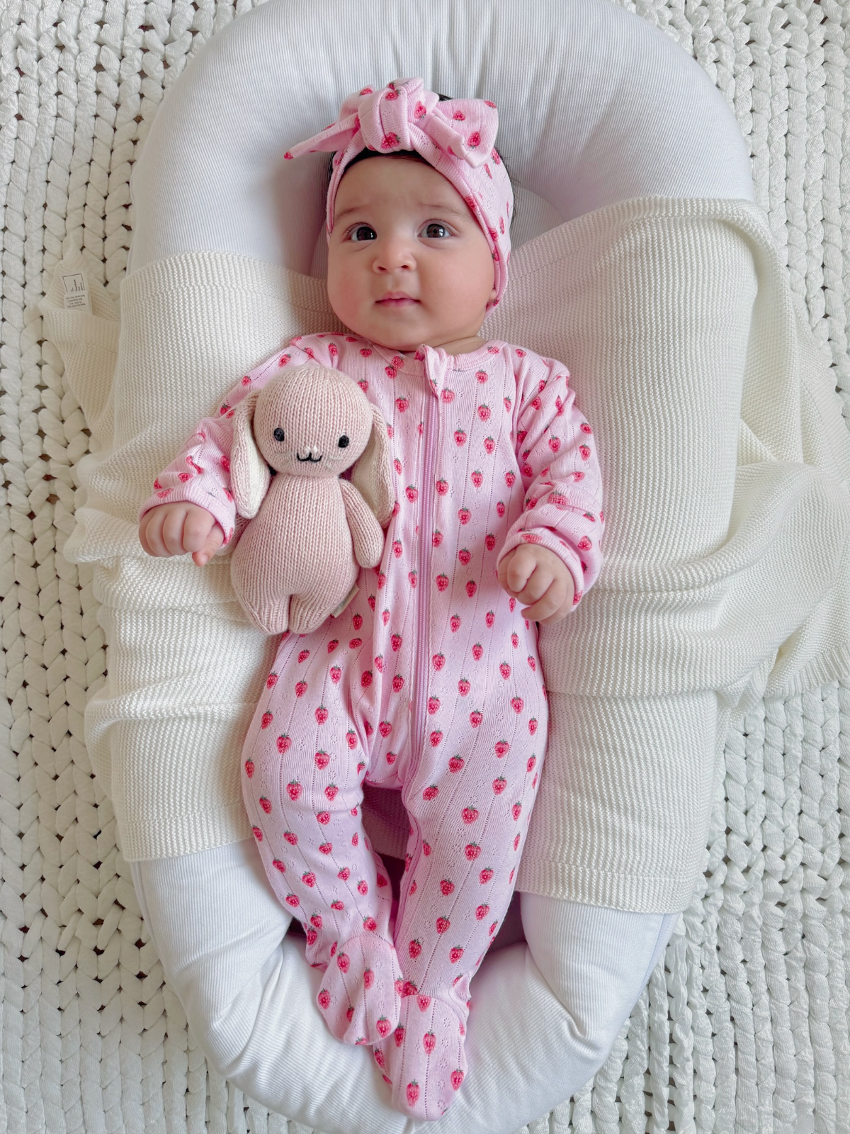 Baby girl in pink strawberry outfit holding a soft bunny, resting on a cozy blanket in a modern bassinet.