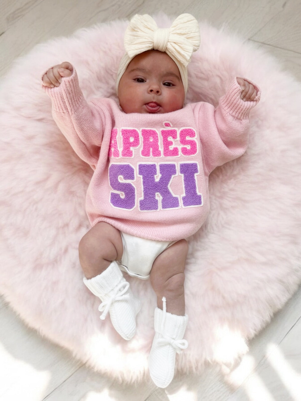 Baby in pink "Après Ski" sweater, lying on a fluffy pink rug, wearing a bow headband and white booties.