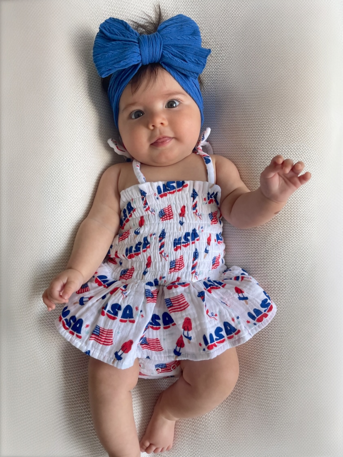 Baby in a patriotic red, white, and blue outfit with a large blue bow, lying on a light background.