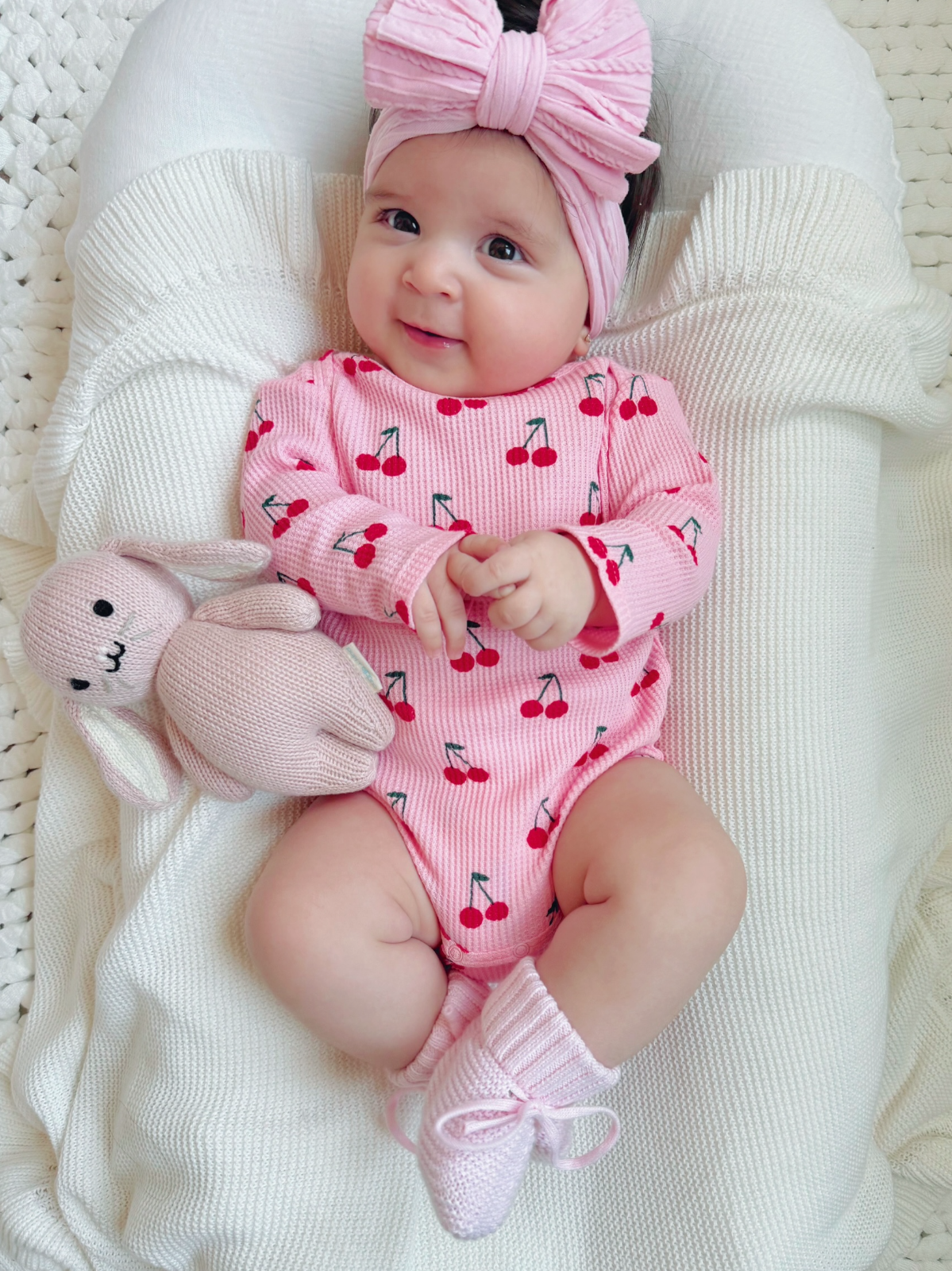 Smiling baby in cherry-patterned onesie and pink headband, holding a soft bunny toy on a cozy blanket.
