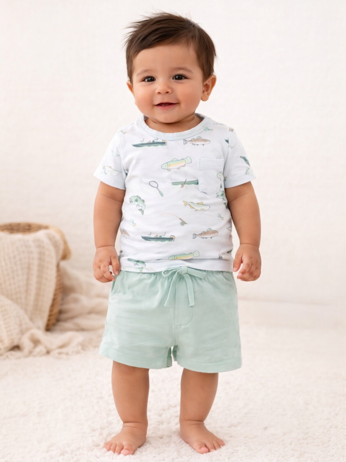 Smiling toddler wearing a fish-patterned shirt and mint shorts, standing on a soft white rug.