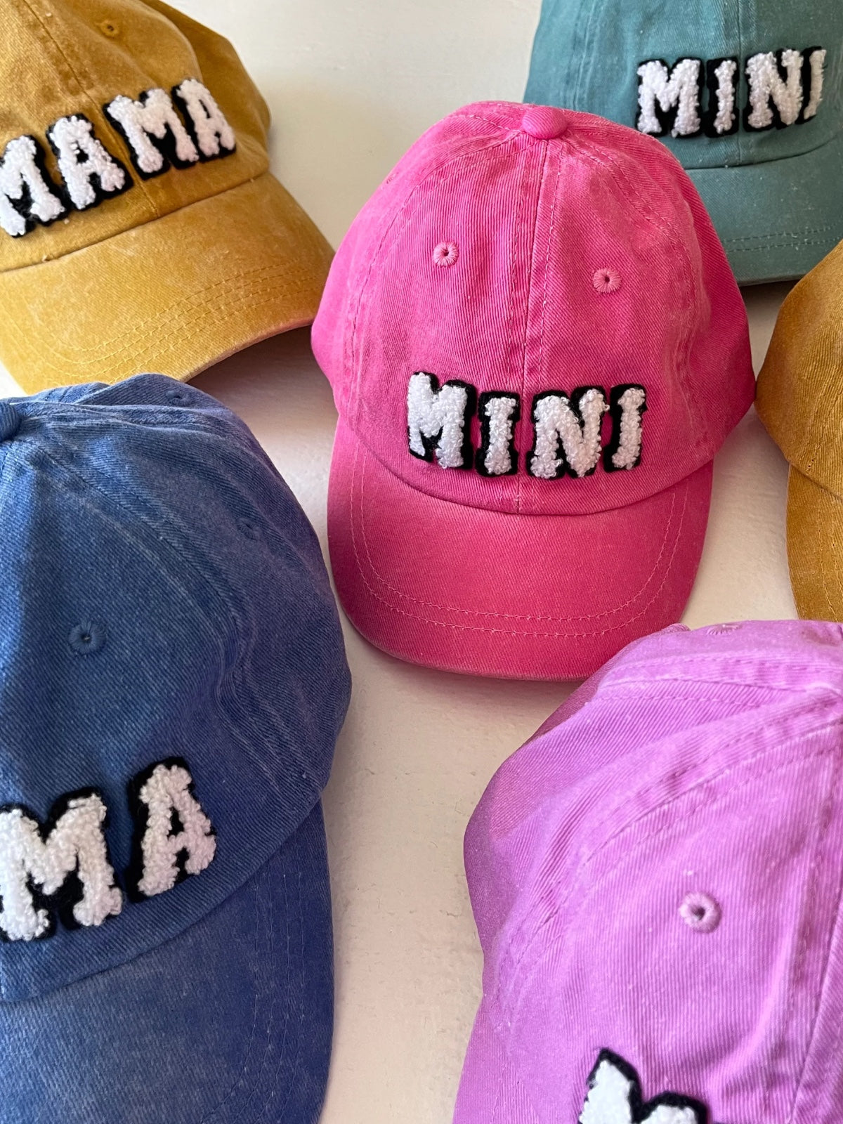Colorful caps with "MAMA" and "MINI" embroidered in fluffy text, arranged on a light surface.