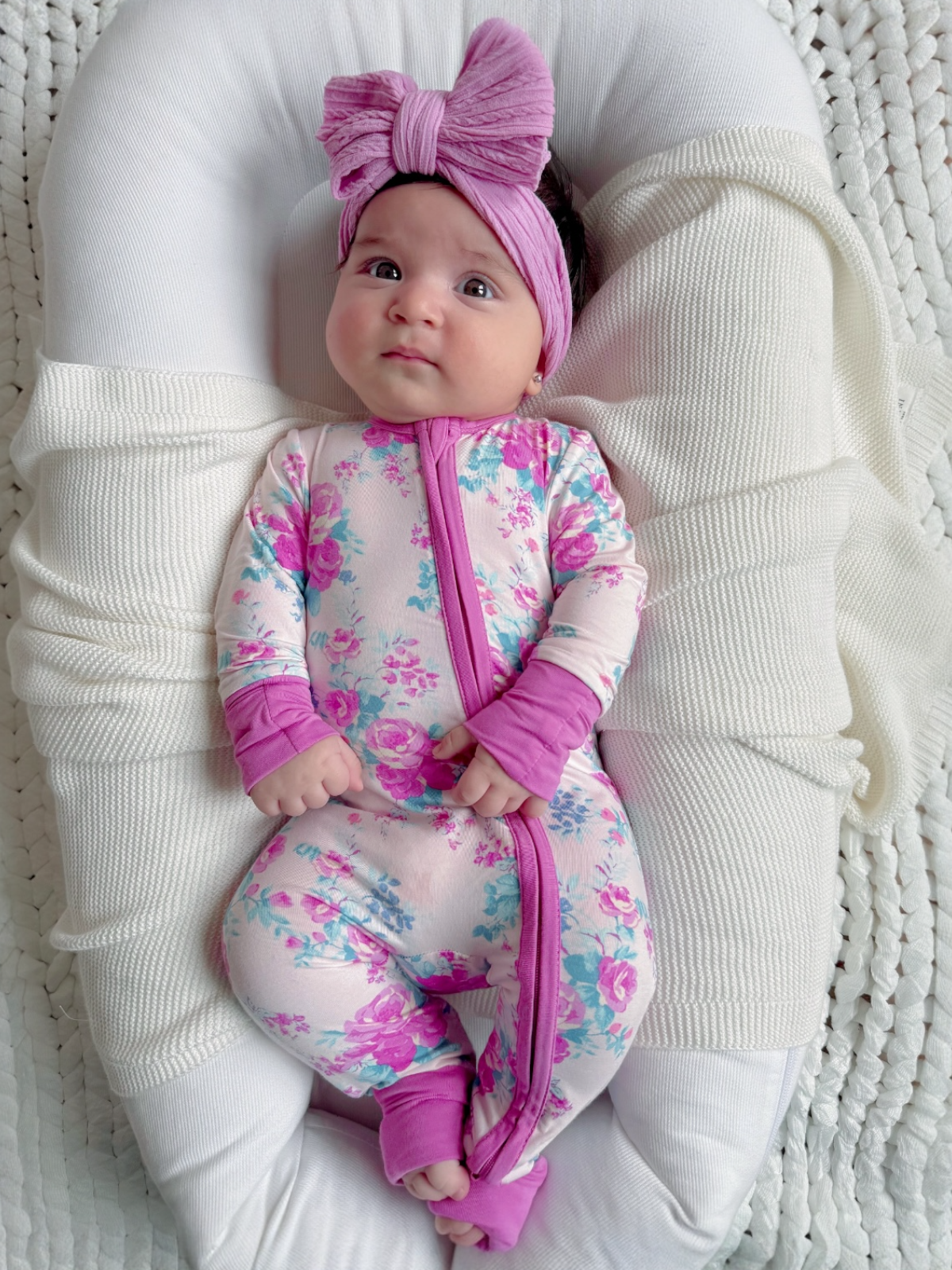 Baby in floral onesie and oversized bow, lying on a soft, textured surface, looking curiously at the camera.