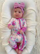 Baby in floral onesie and pink bow headband, resting comfortably on a soft blanket.
