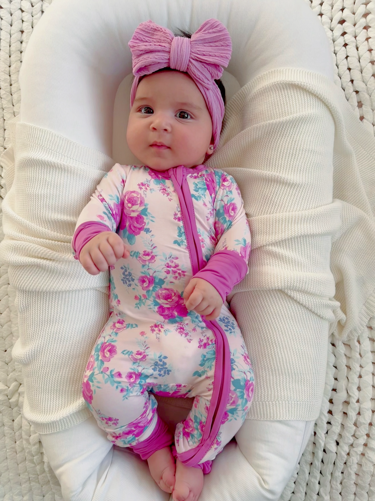Baby in floral onesie and pink bow headband, resting comfortably on a soft blanket.