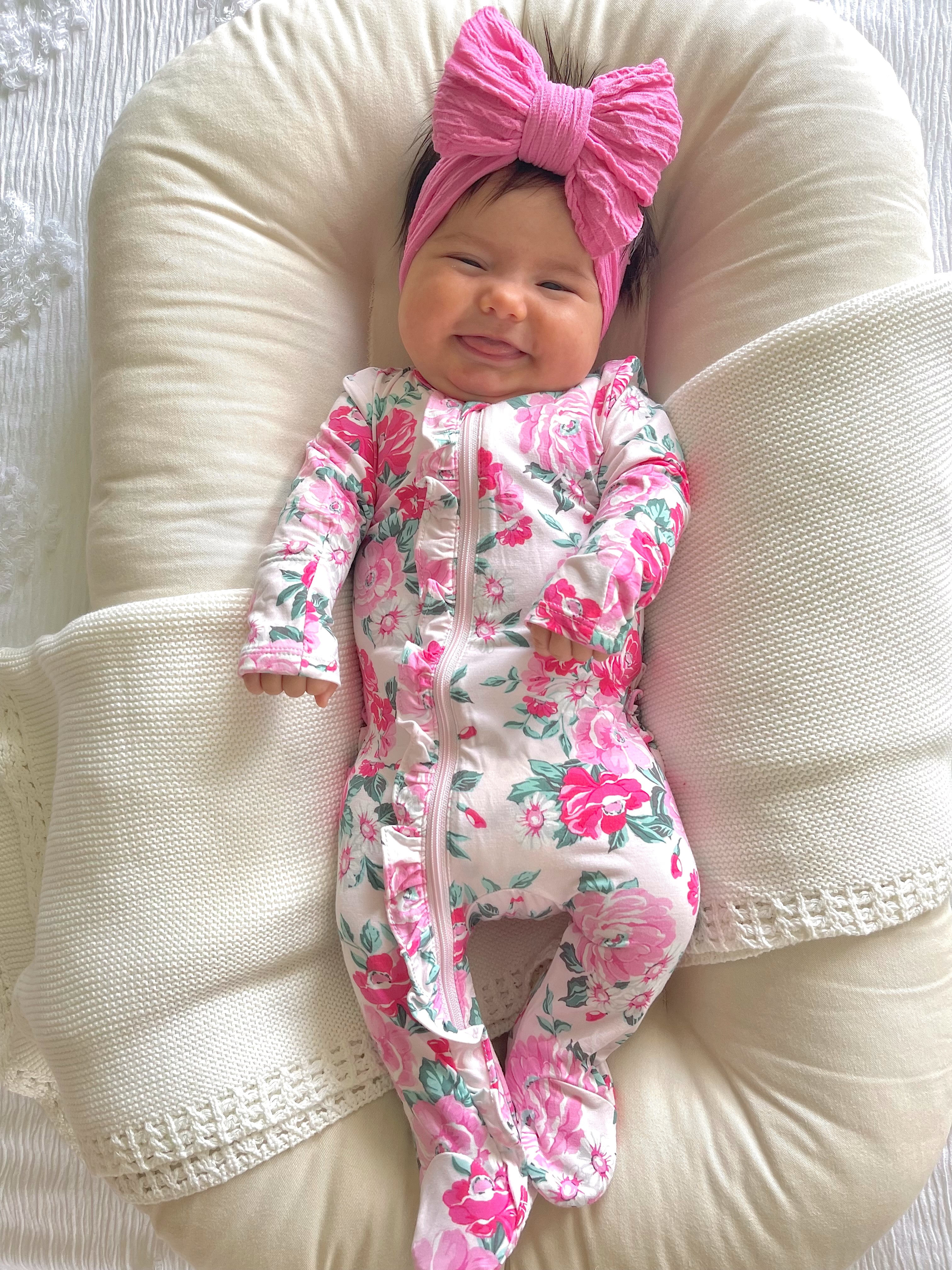 Smiling baby girl in floral onesie and pink bow, resting comfortably on a soft pillow.