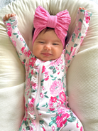 Smiling baby girl in floral onesie with pink bow, stretching happily on a plush pillow.