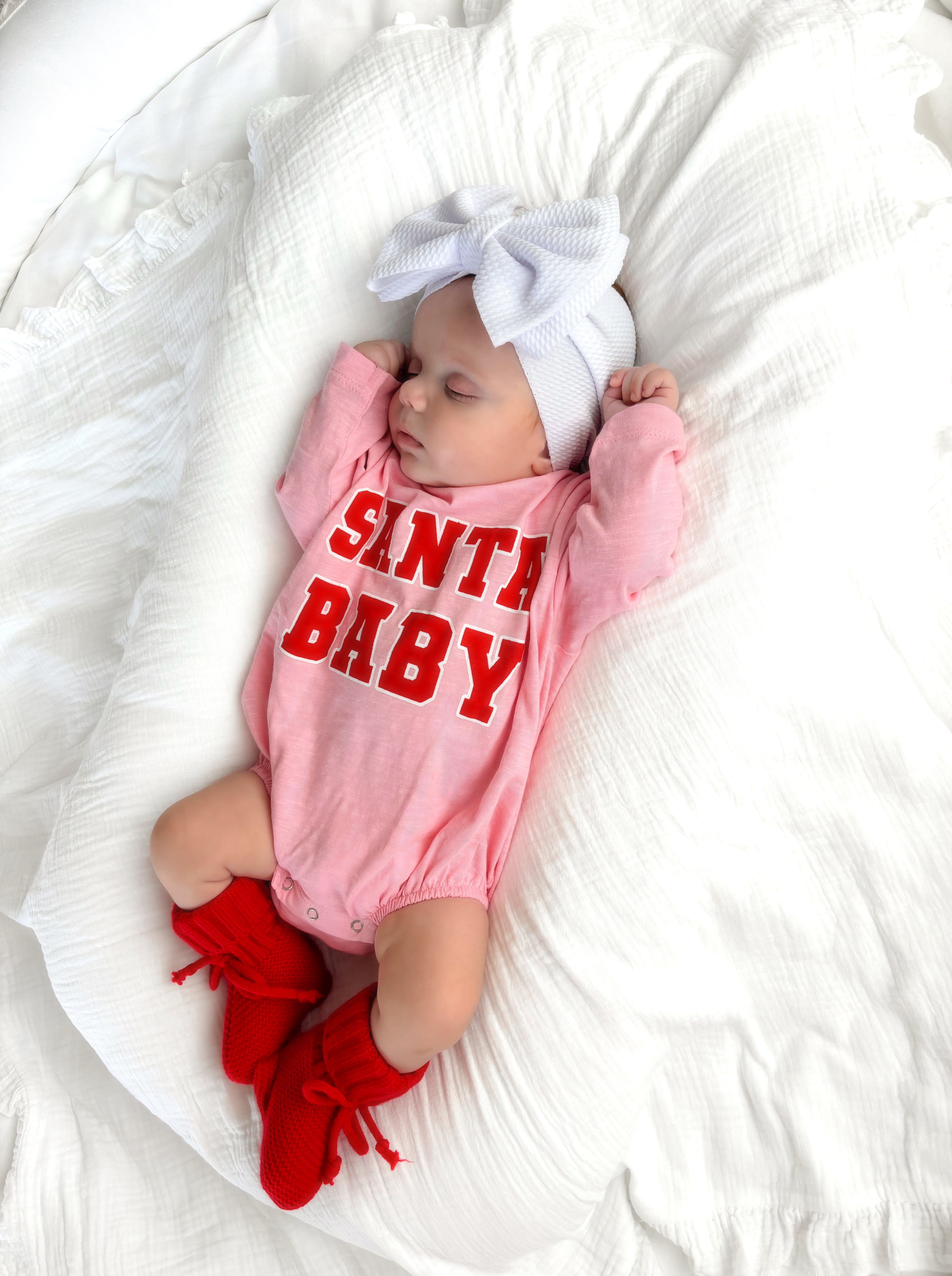 Sleeping baby in pink onesie with "Santa Baby" text and red booties, on a white blanket.