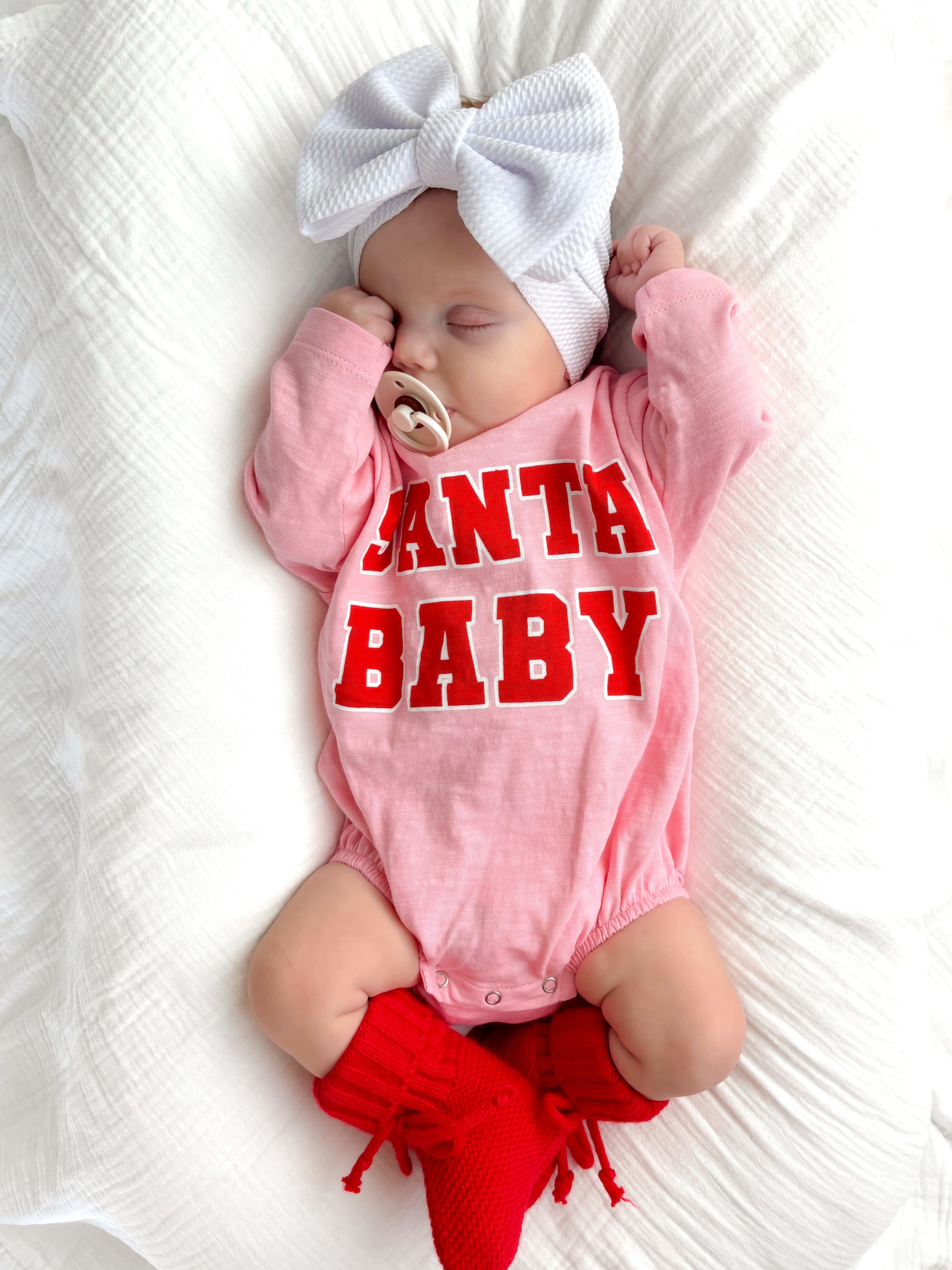 Sleeping baby in pink onesie with "Santa Baby" text, white headband, and red knitted socks on a white blanket.