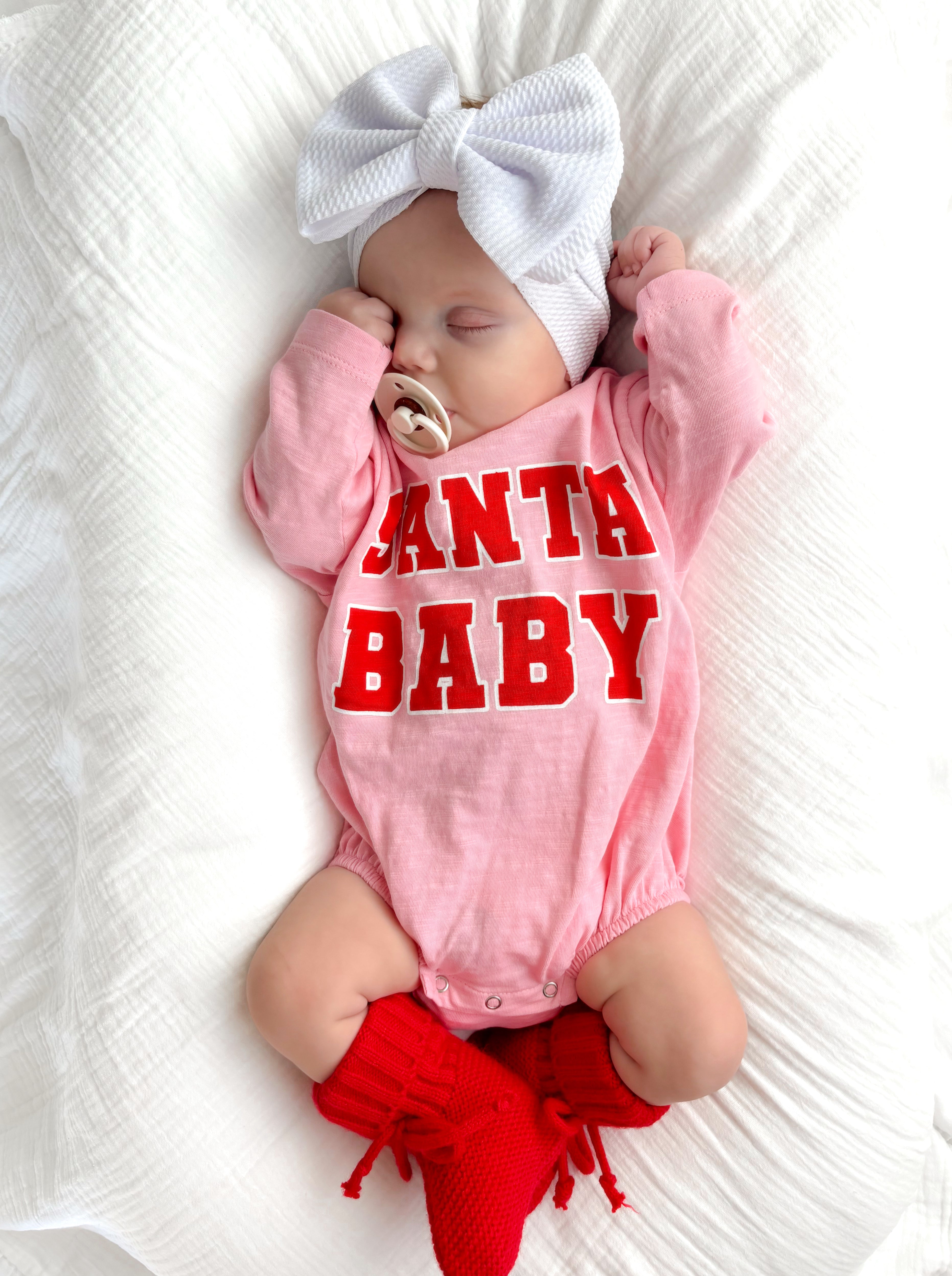 Sleeping baby in pink onesie with "Santa Baby" text, white headband, and red knitted socks on a white blanket.