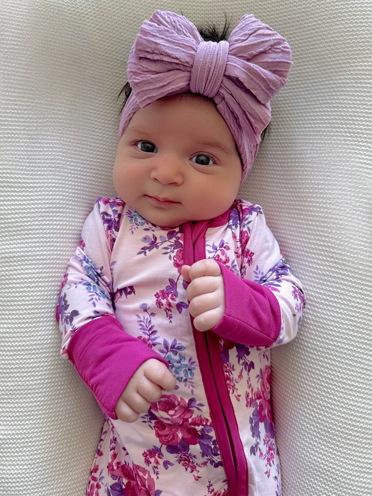 Smiling baby girl wearing a floral outfit and a large purple headband, lying on a textured white surface.