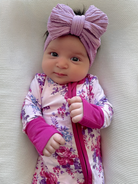 Smiling baby girl wearing a floral outfit and a large purple headband, lying on a textured white surface.