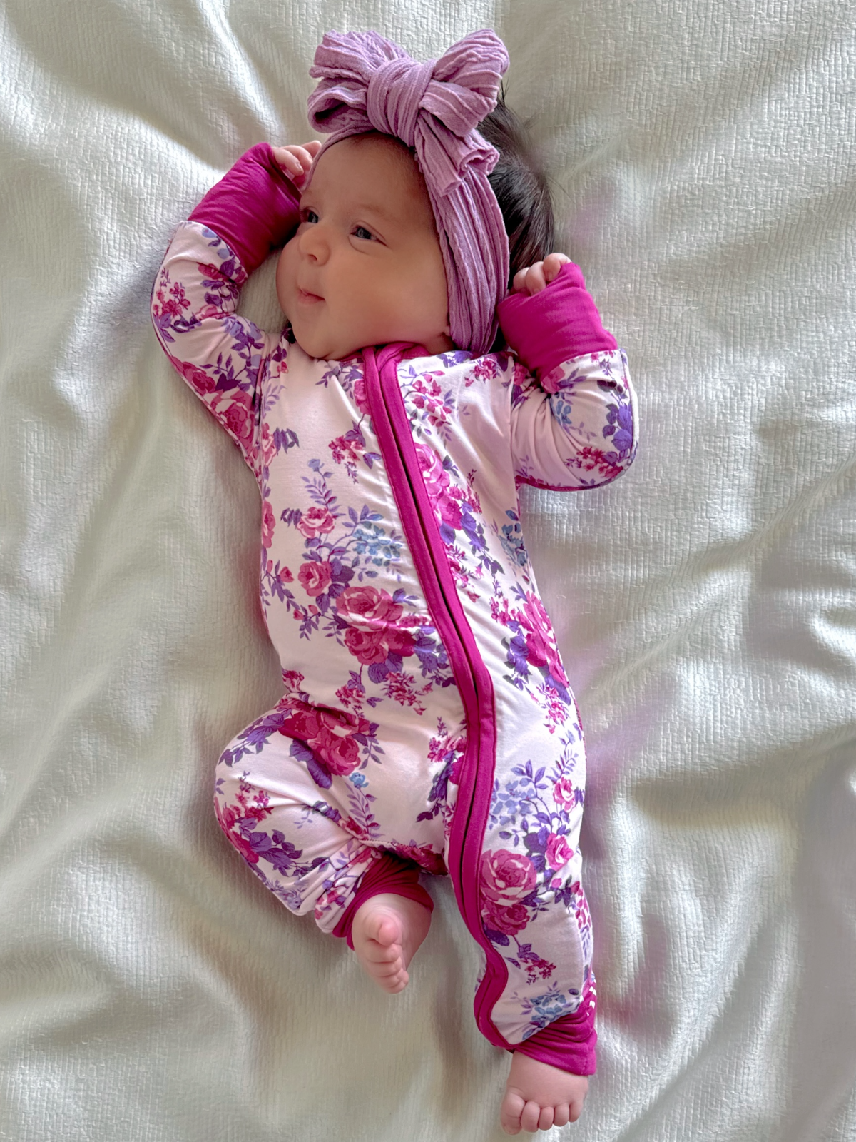 Baby in floral onesie and headband, lying on a soft blanket, smiling and looking playfully at the camera.