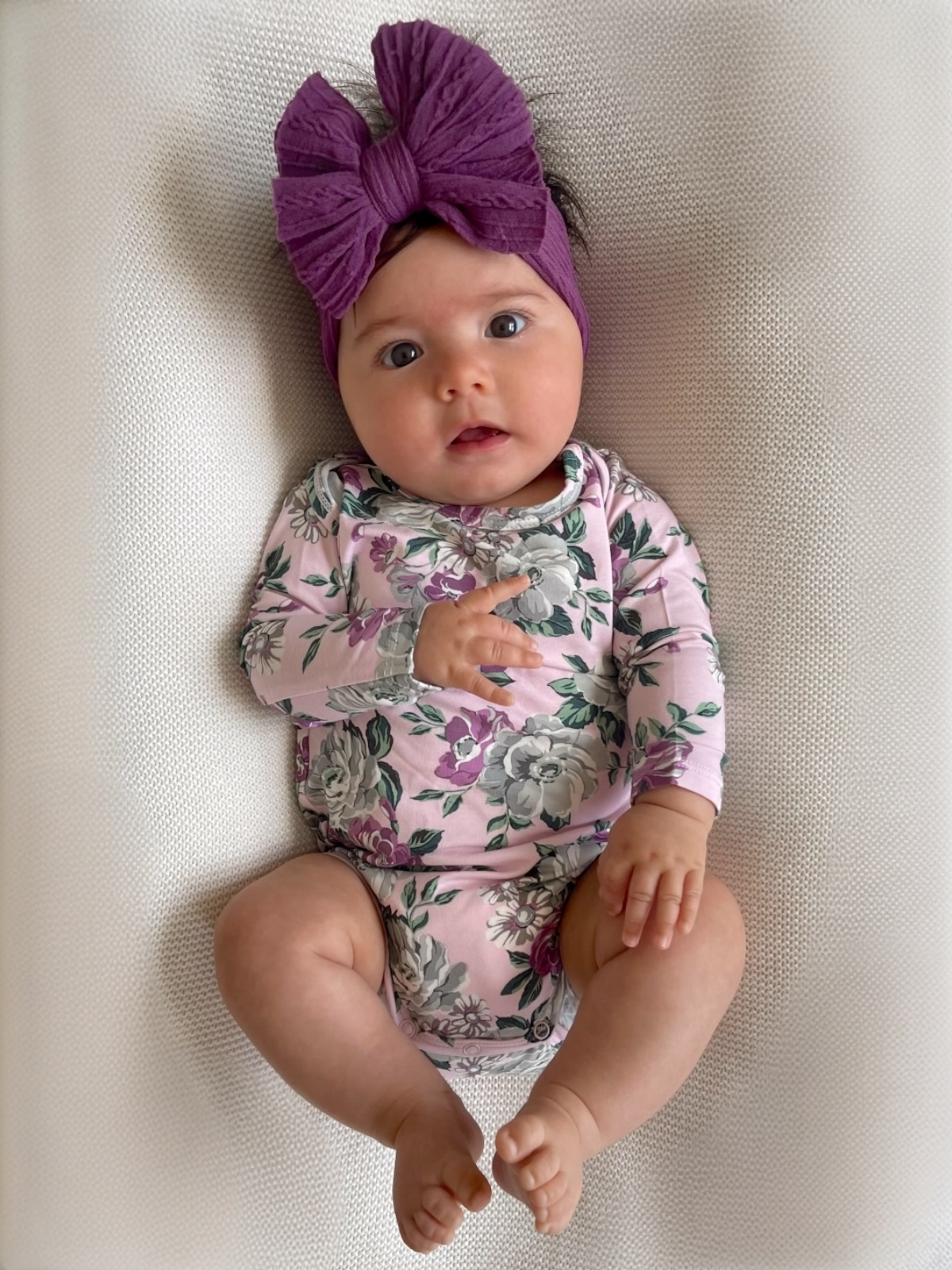 Infant girl lying on a blanket, wearing a floral outfit and a large purple headband, with a curious expression.