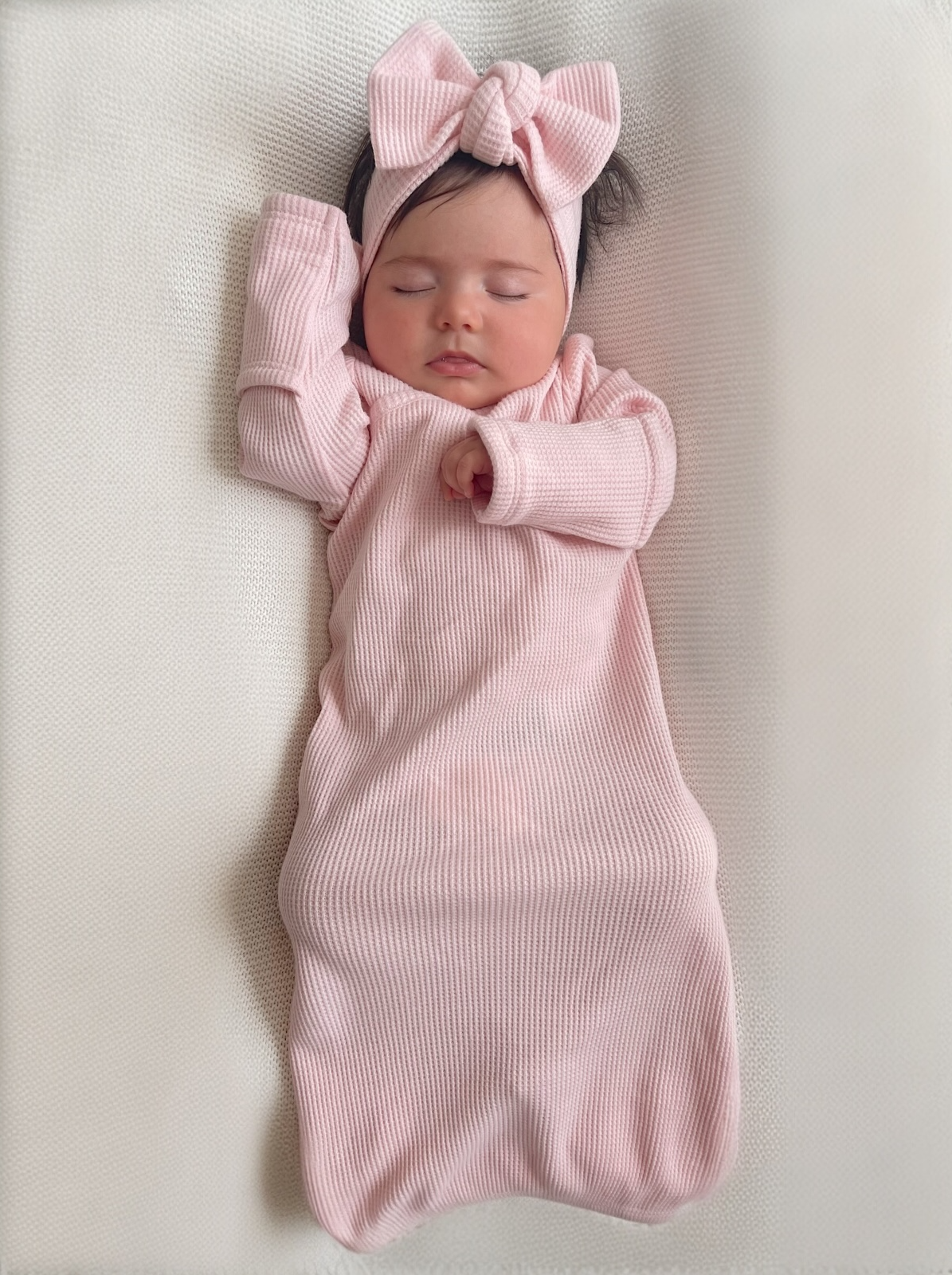 Infant sleeping peacefully in a pink outfit and headband, resting on a textured white surface.