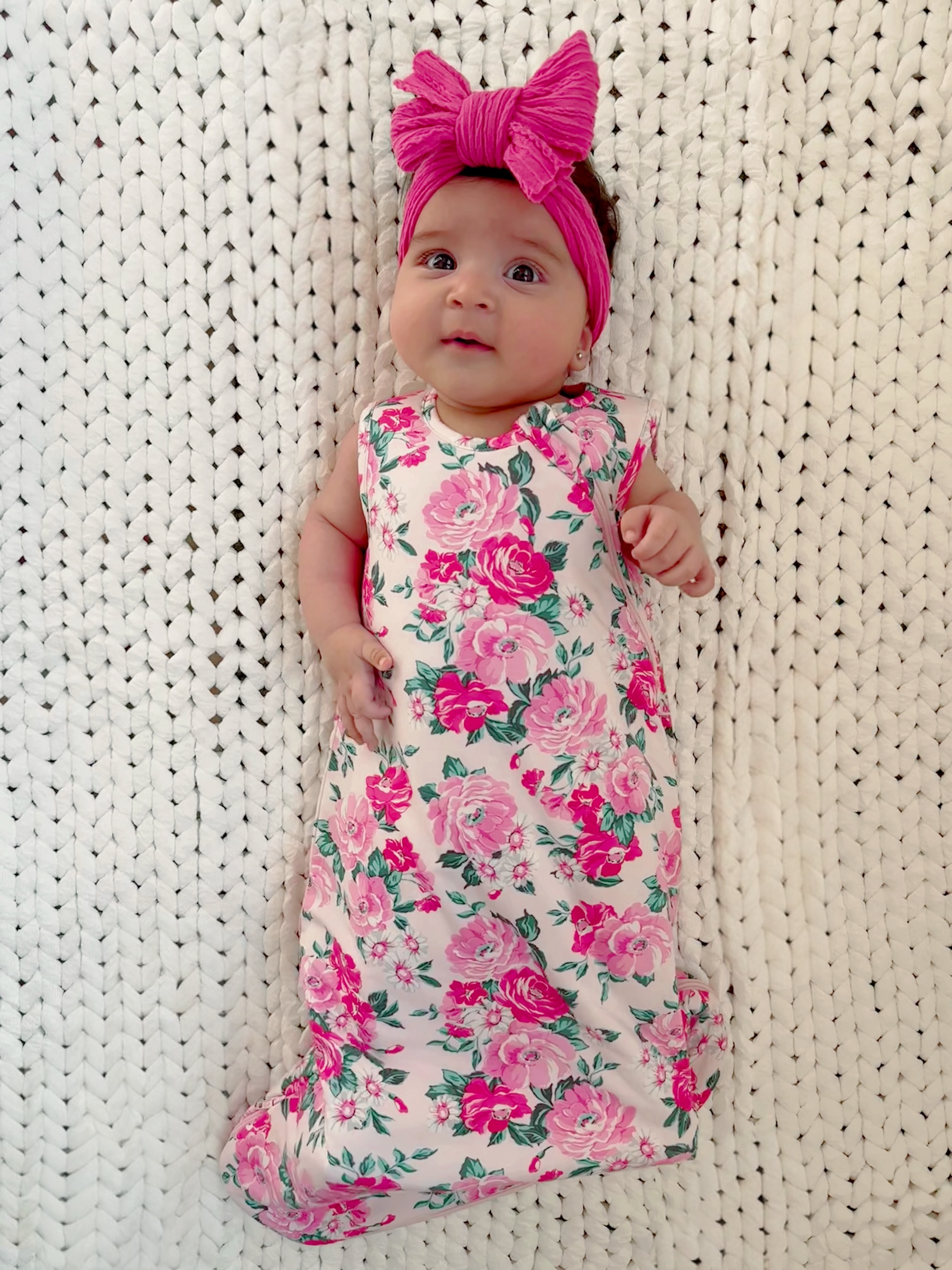 Baby girl in a floral dress with a pink bow, lying on a white knitted blanket, smiling at the camera.
