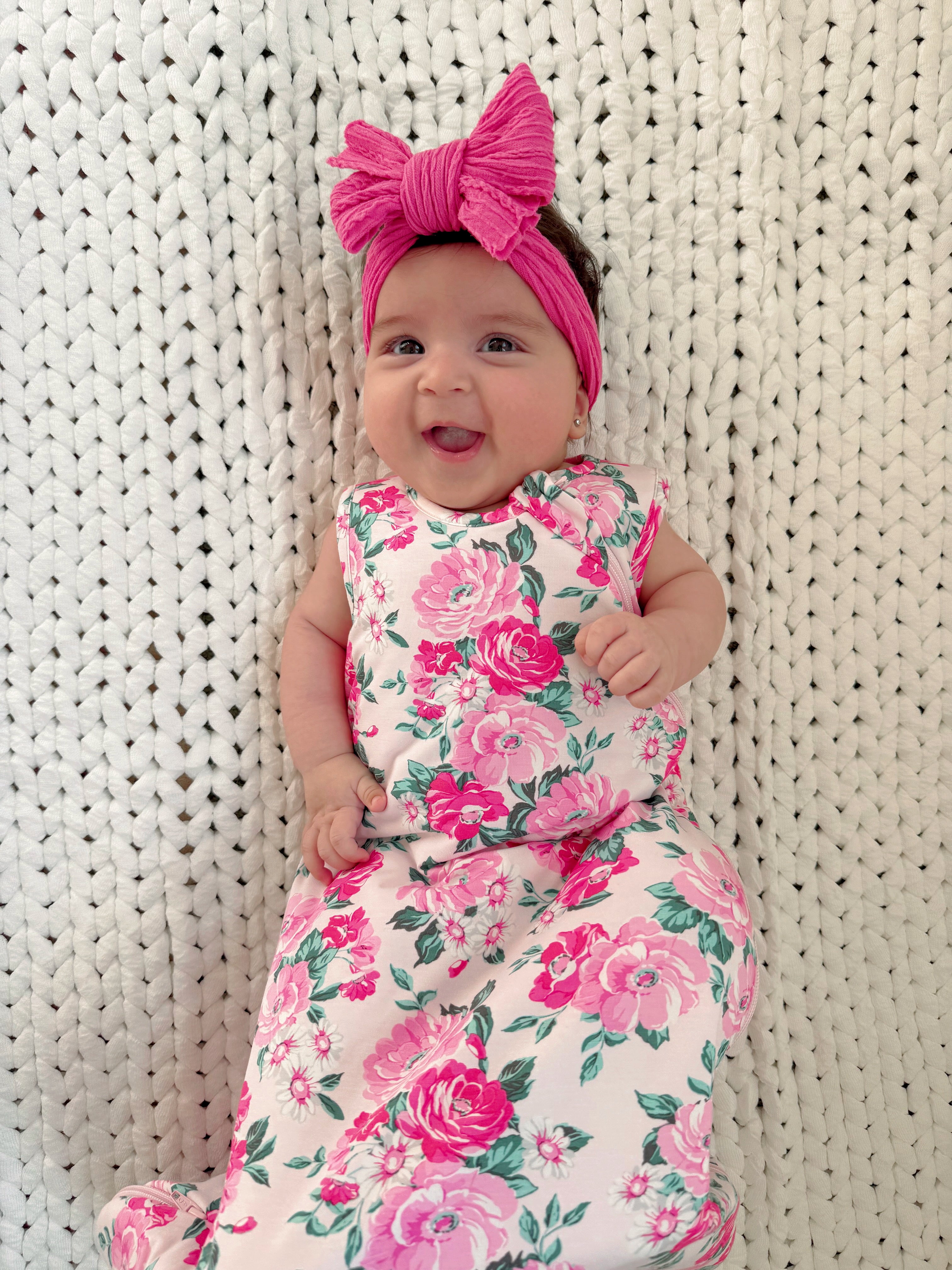 Smiling baby girl in floral dress and pink headband, lying on a textured white blanket.