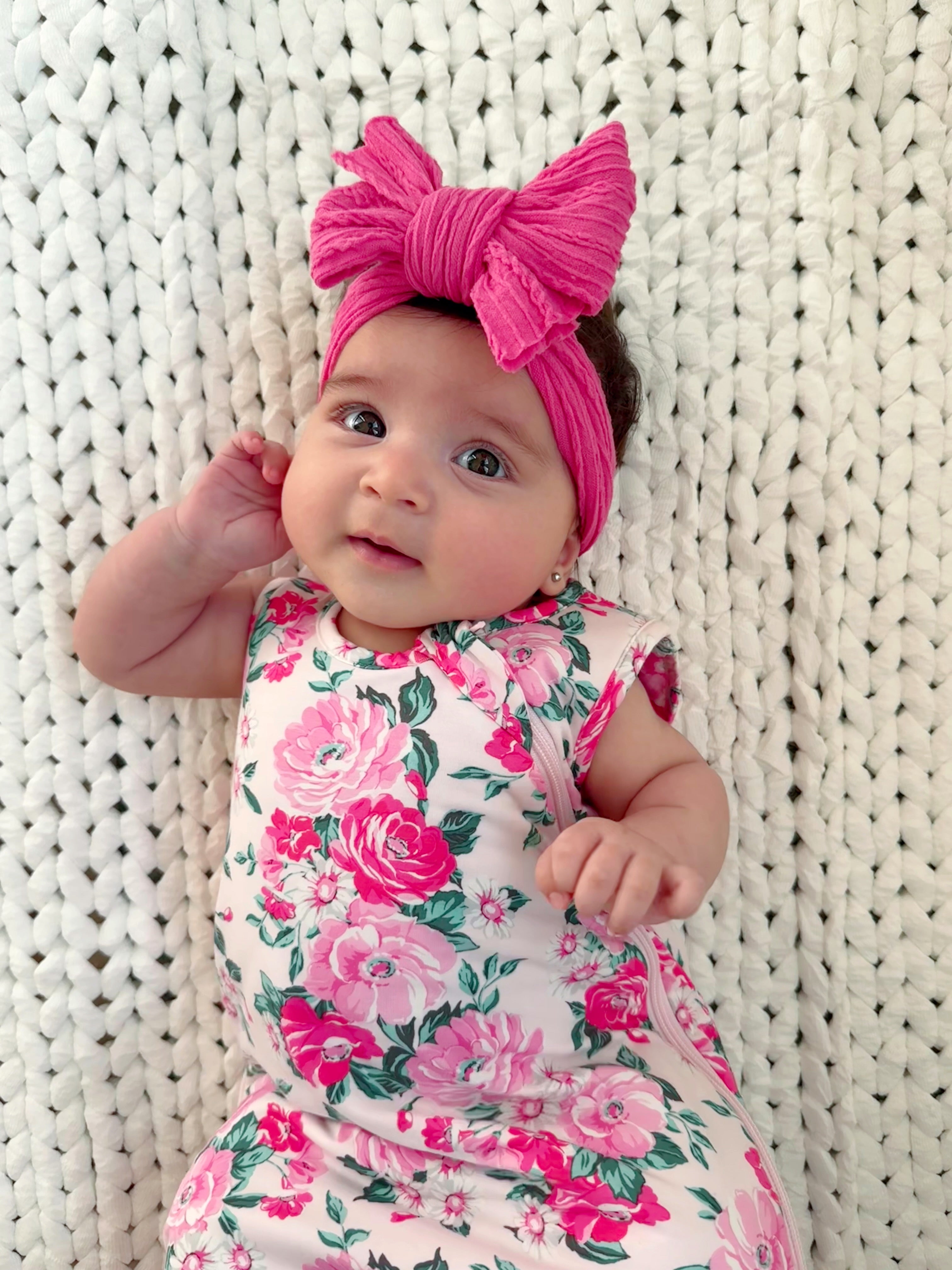 Infant with a pink bow headband, wearing a floral dress, posed on a cozy knitted blanket, smiling at the camera.