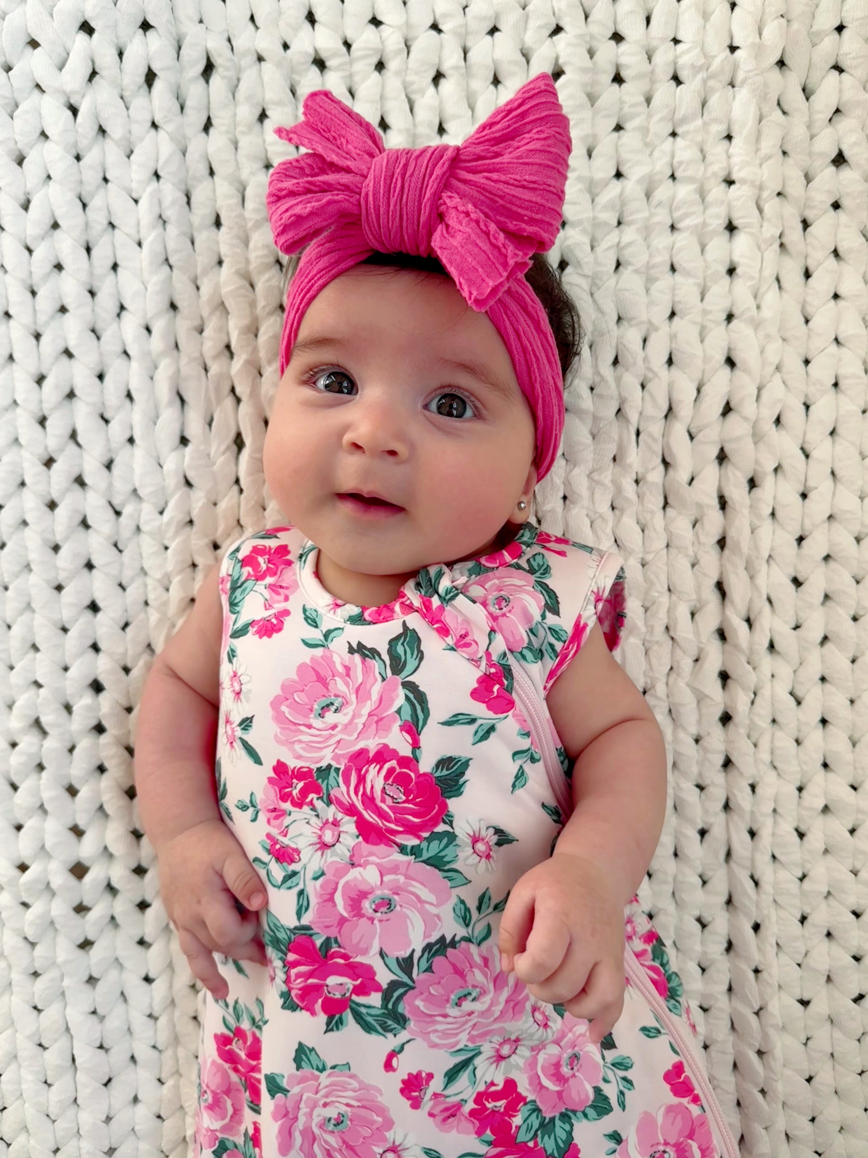 Baby girl in a floral dress and pink bow headband, against a cozy, knitted background, smiling sweetly.