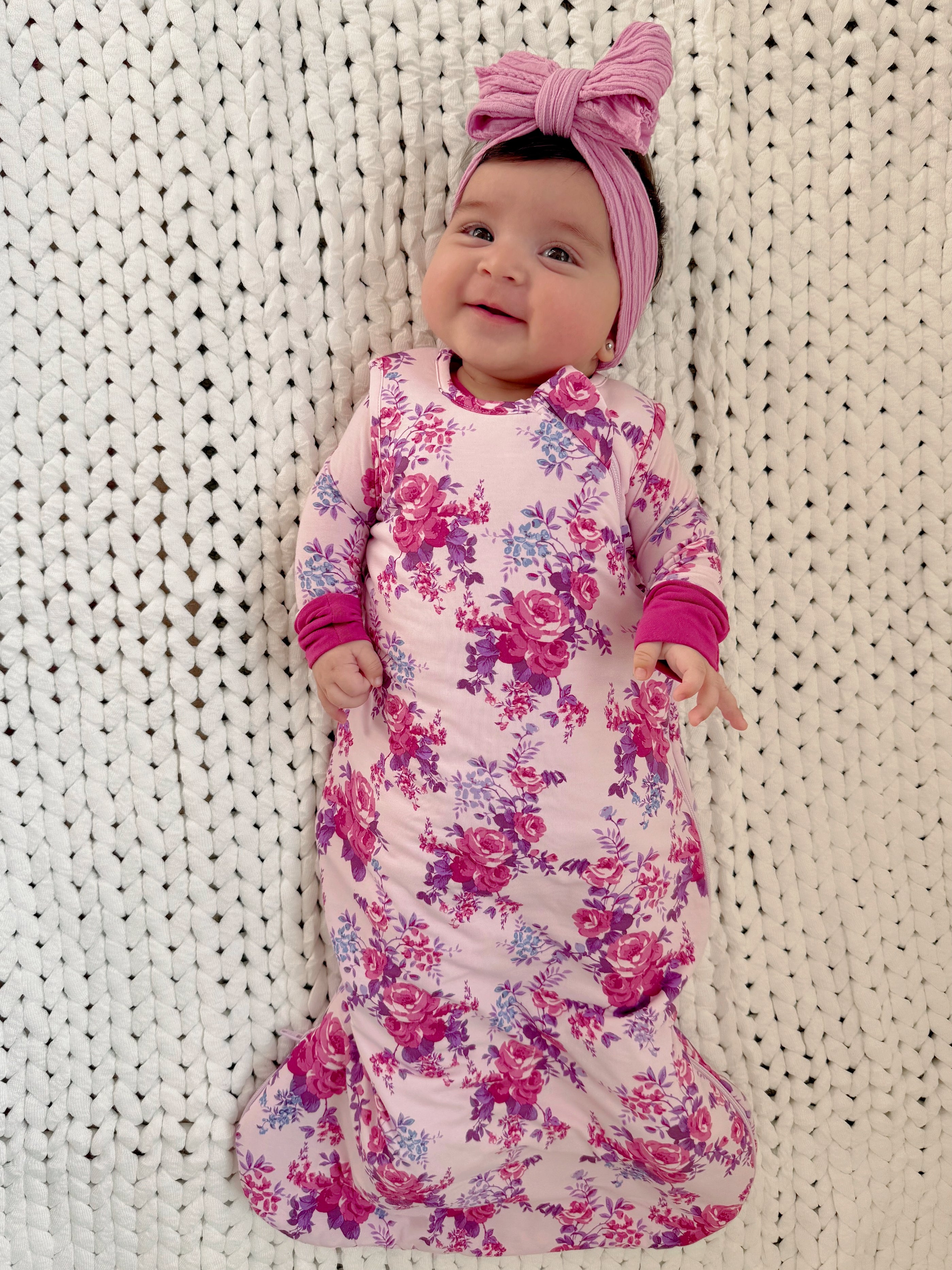 Smiling baby in floral swaddle and pink headband, lying on a textured white blanket.