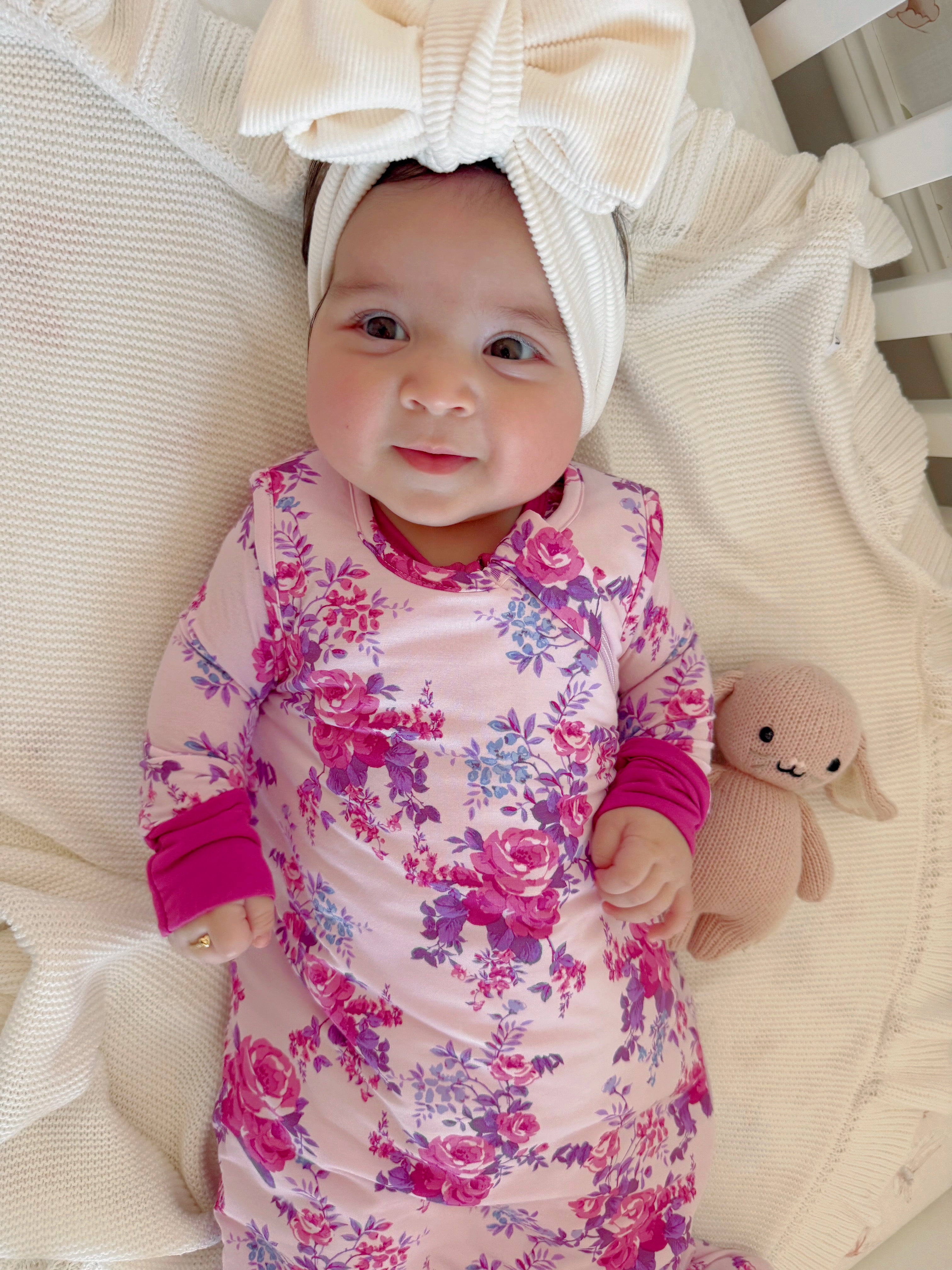 Smiling baby in floral dress with large bow headband and stuffed bunny on a cozy blanket.