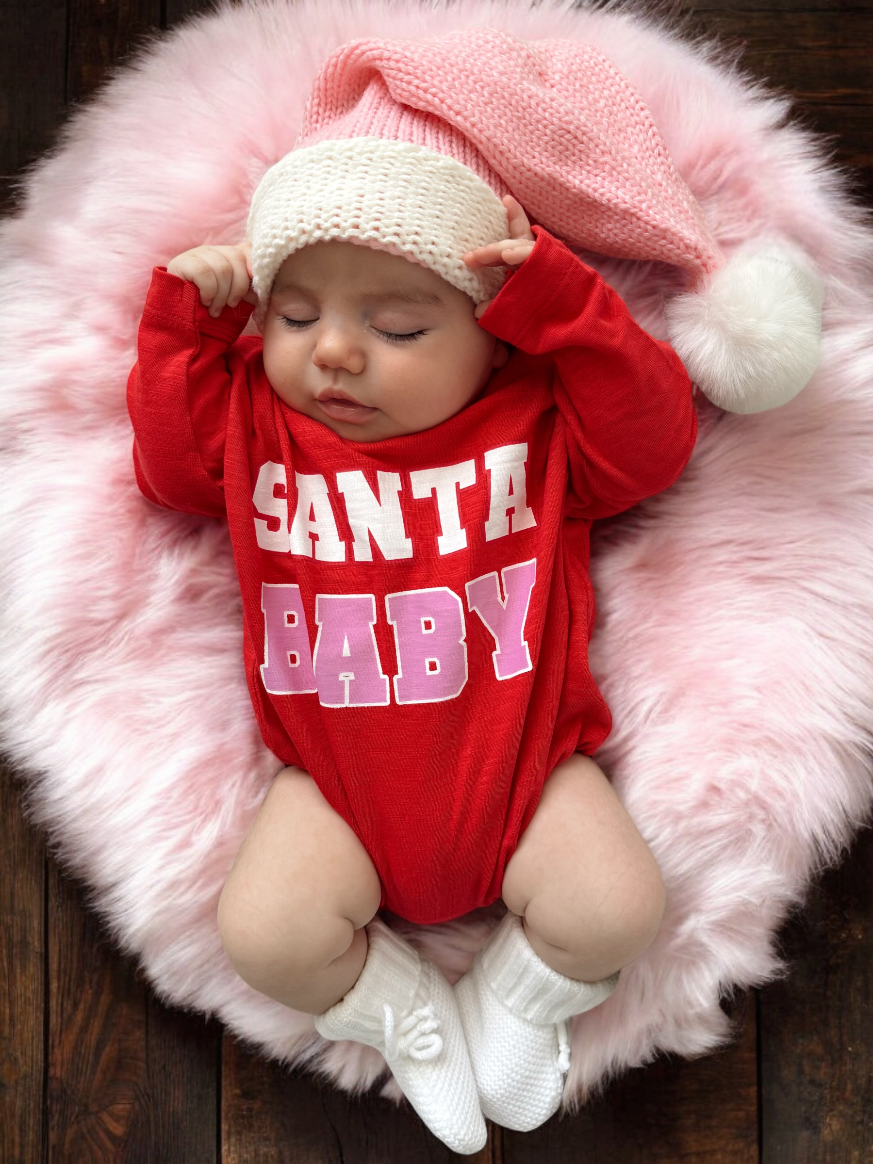 Sleeping baby in a red "Santa Baby" onesie, wearing a pink knitted hat, on a fluffy pink blanket.
