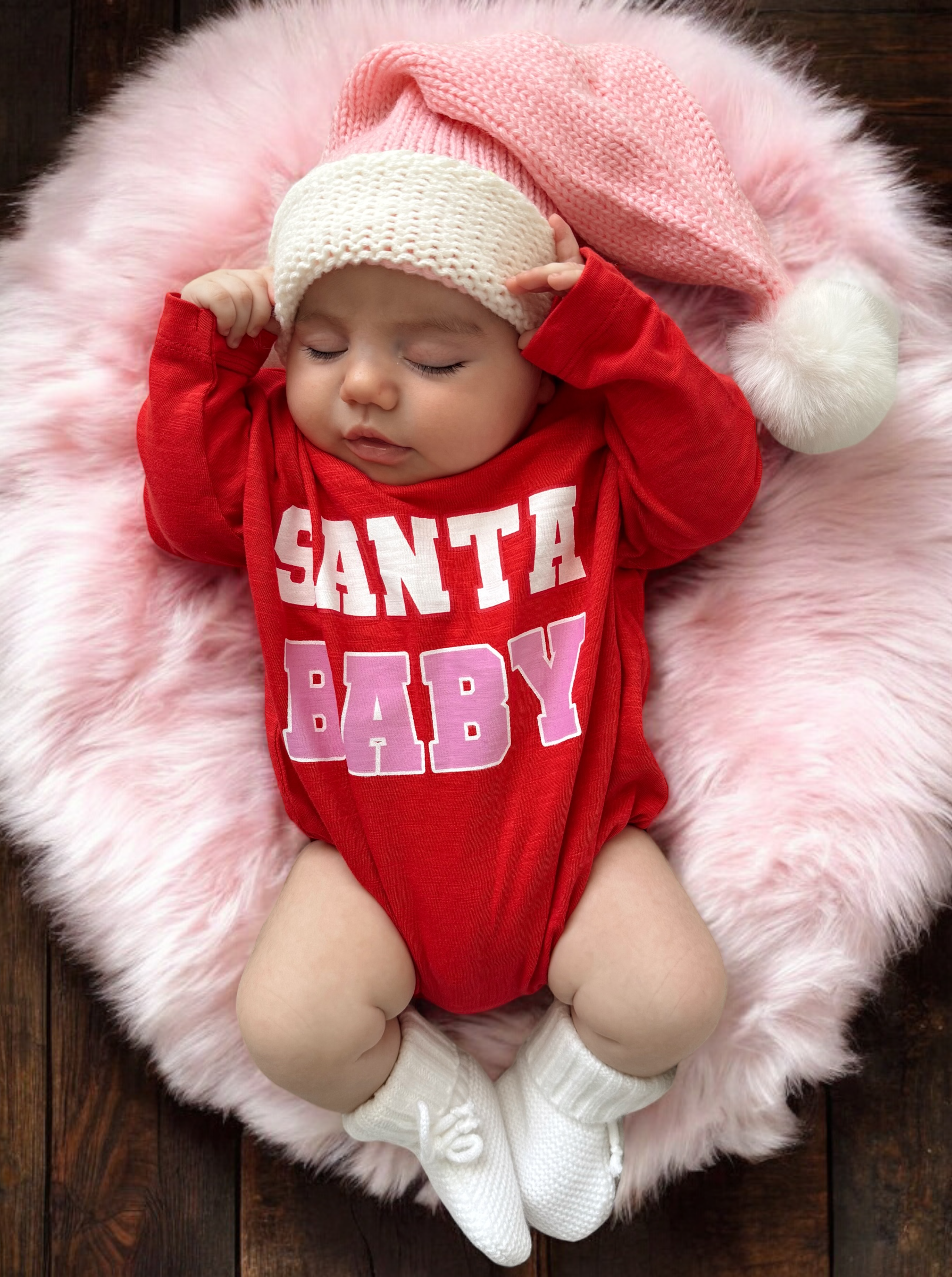 Sleeping baby in a red "Santa Baby" onesie, wearing a pink knitted hat, on a fluffy pink blanket.