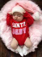 Sleeping baby in a red "Santa Baby" onesie, wearing a pink knitted hat, on a fluffy pink blanket.
