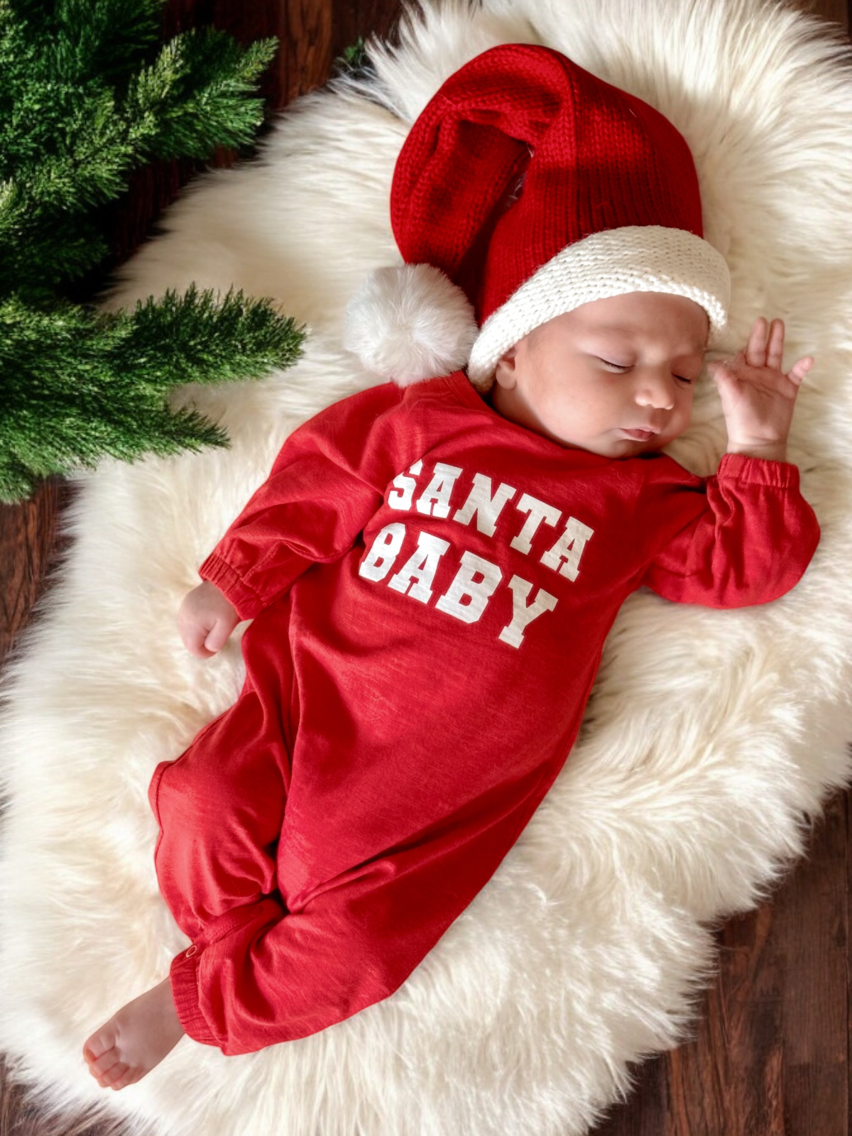 Sleeping baby in a red "Santa Baby" onesie and a festive hat, relaxing on a fluffy white blanket with greenery.