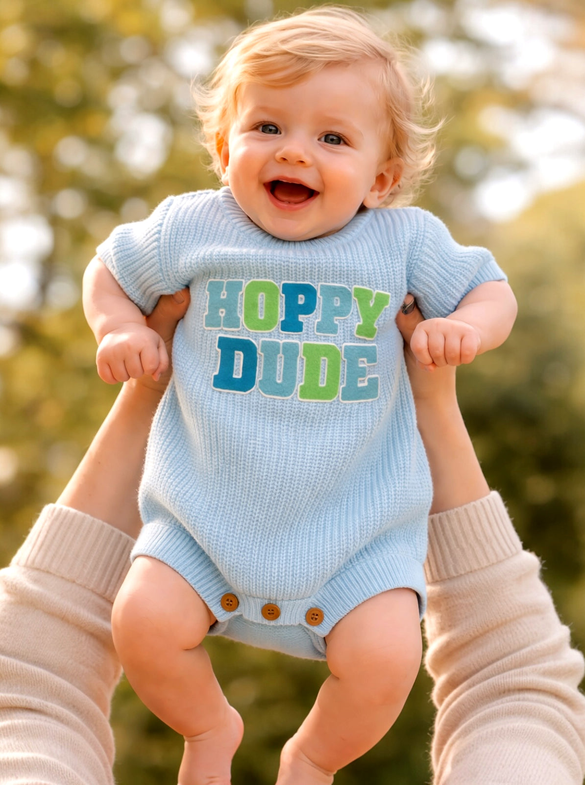 Smiling baby in a blue onesie with "Hoppy Dude" text, held by a person against a blurred outdoor background.