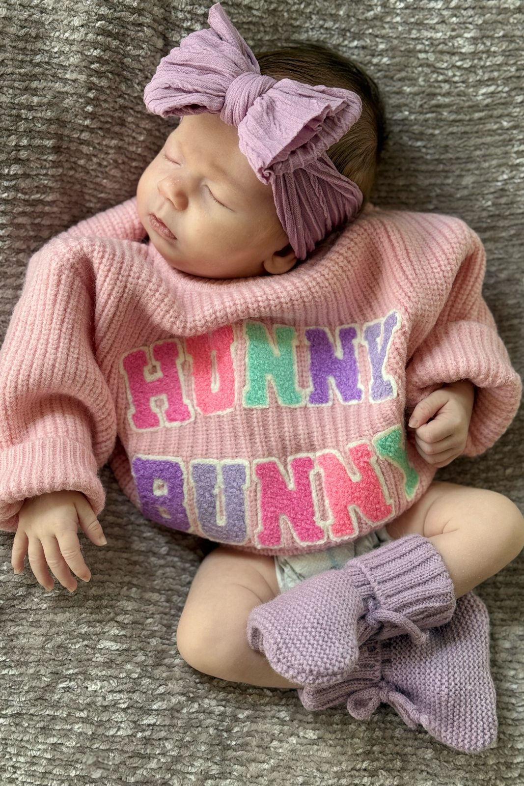 Baby girl in pink "Hunny Bunny" sweater and purple headband, resting on a cozy gray blanket.