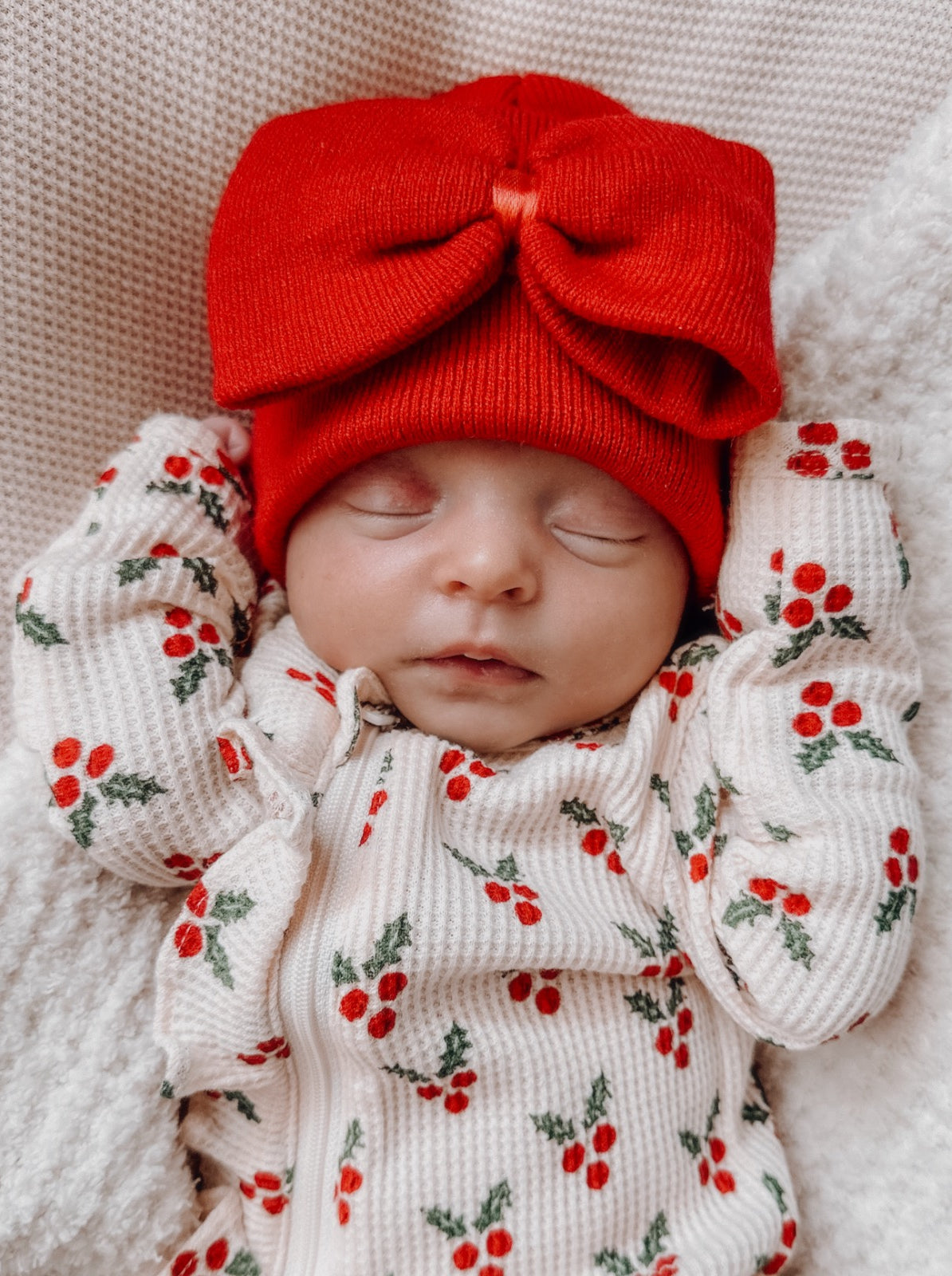 Sleeping baby wearing a red bow hat and a cherry-patterned outfit, resting on a soft white blanket.