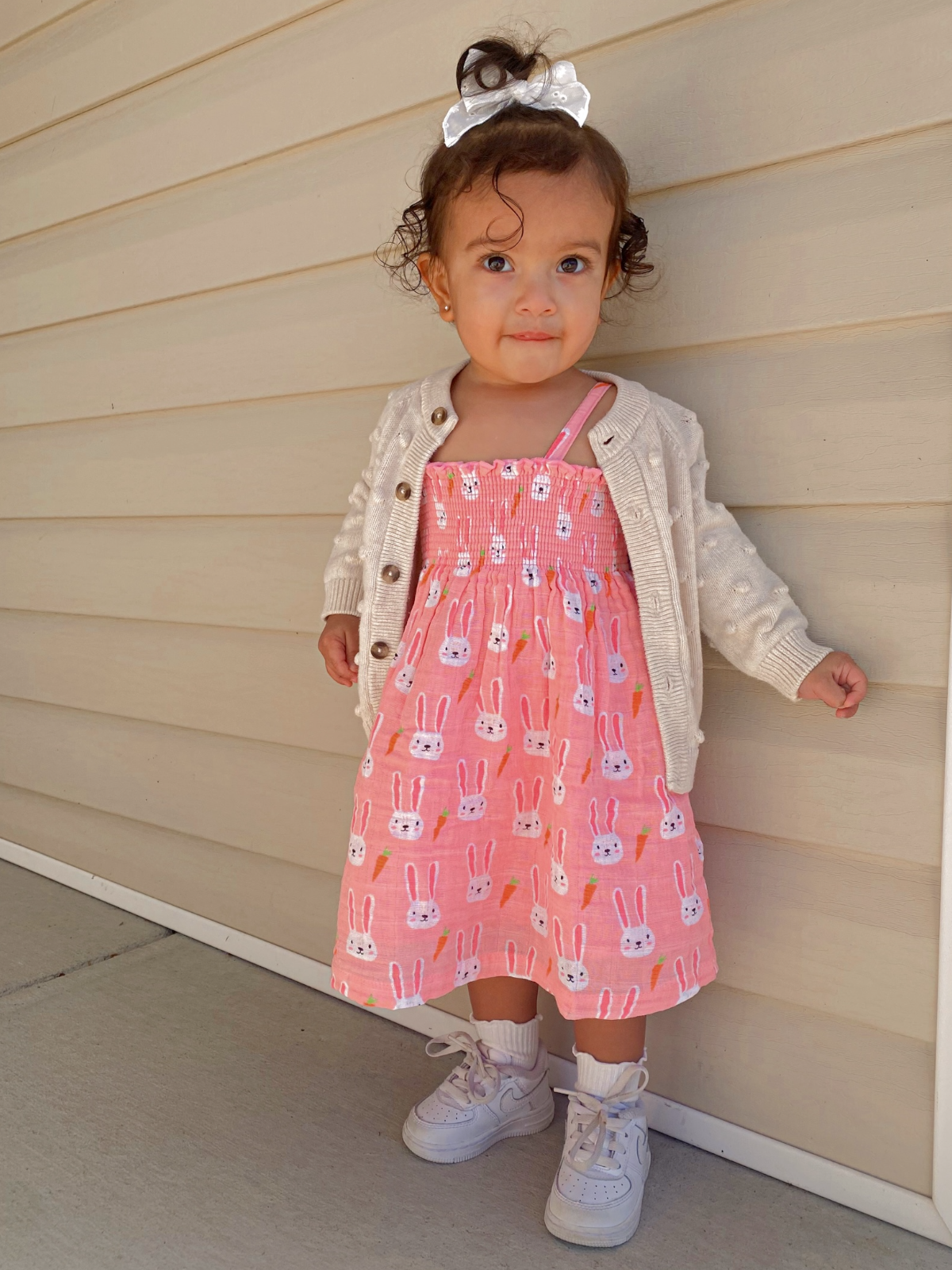 Toddler in a pink bunny-printed dress and cardigan, standing against a light wall with a playful expression.