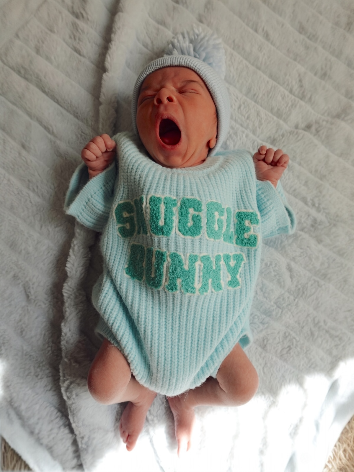 A yawning baby in a light blue outfit with "Snuggle Bunny" text, lying on a soft, textured blanket.