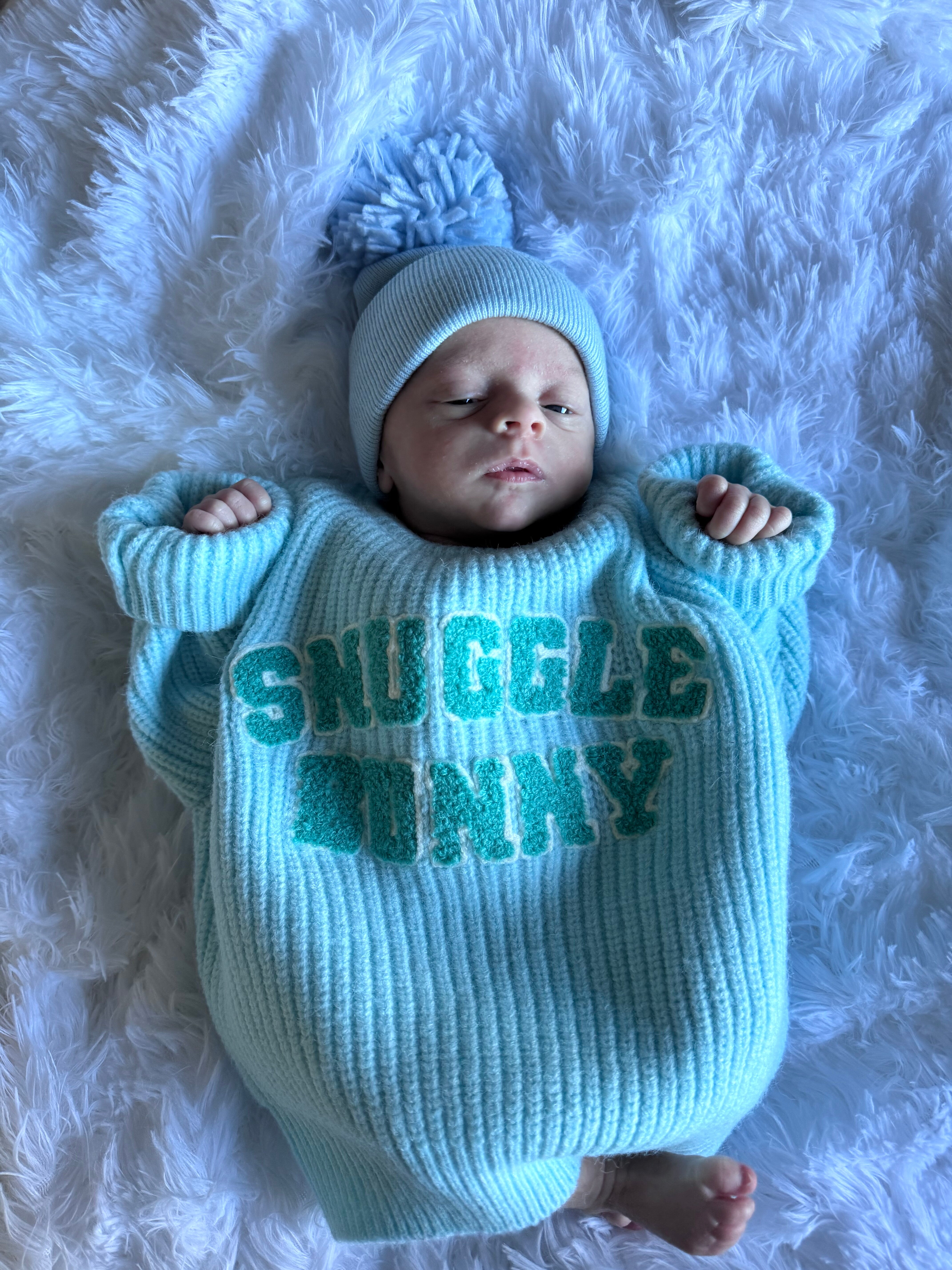 Baby in a light blue oversized sweater with "Snuggle Bunny" text, lying on a fluffy white surface.