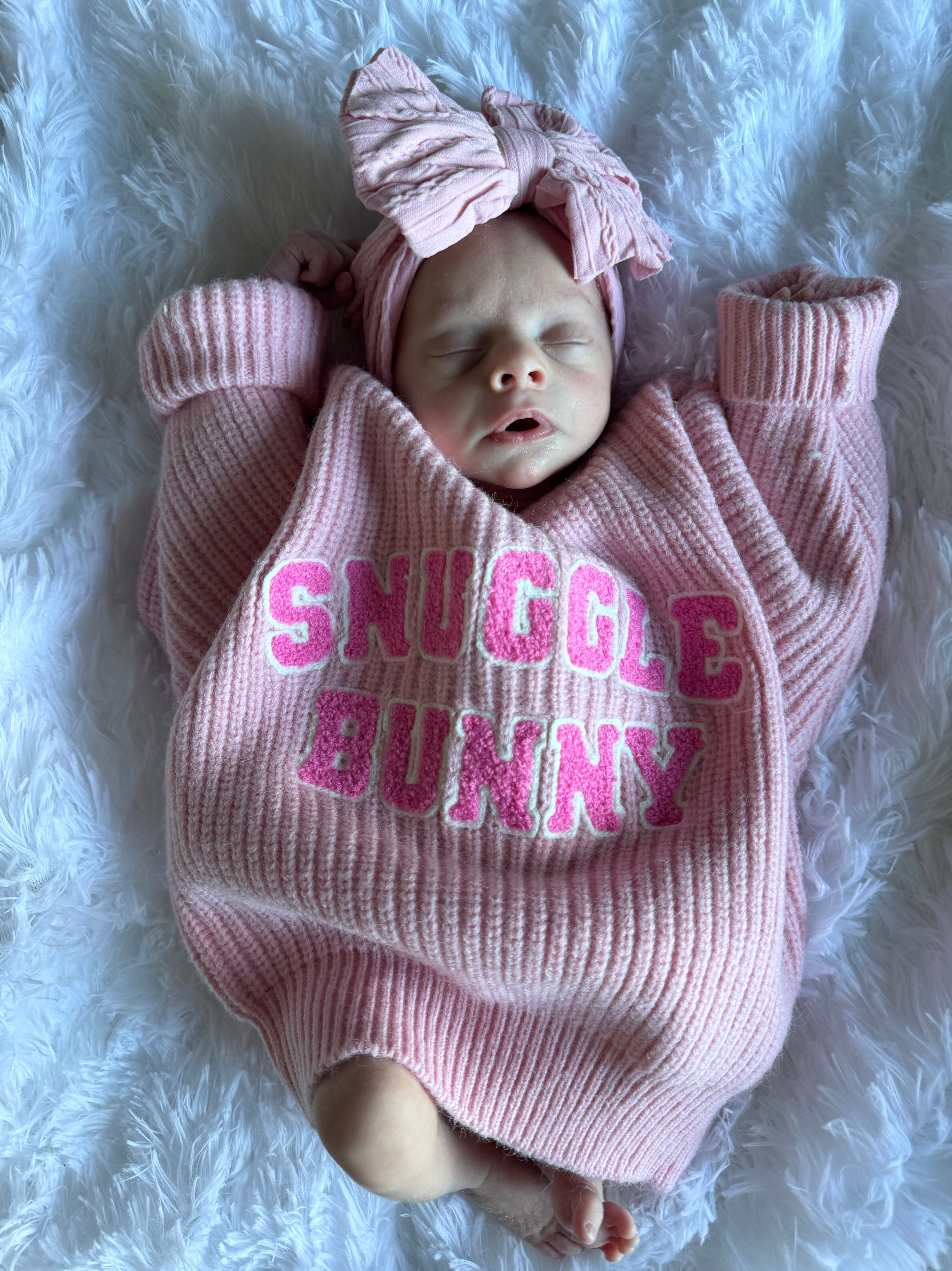 A sleeping baby in a pink sweater with "Snuggle Bunny" text, wearing a matching headband, on a fluffy white blanket.