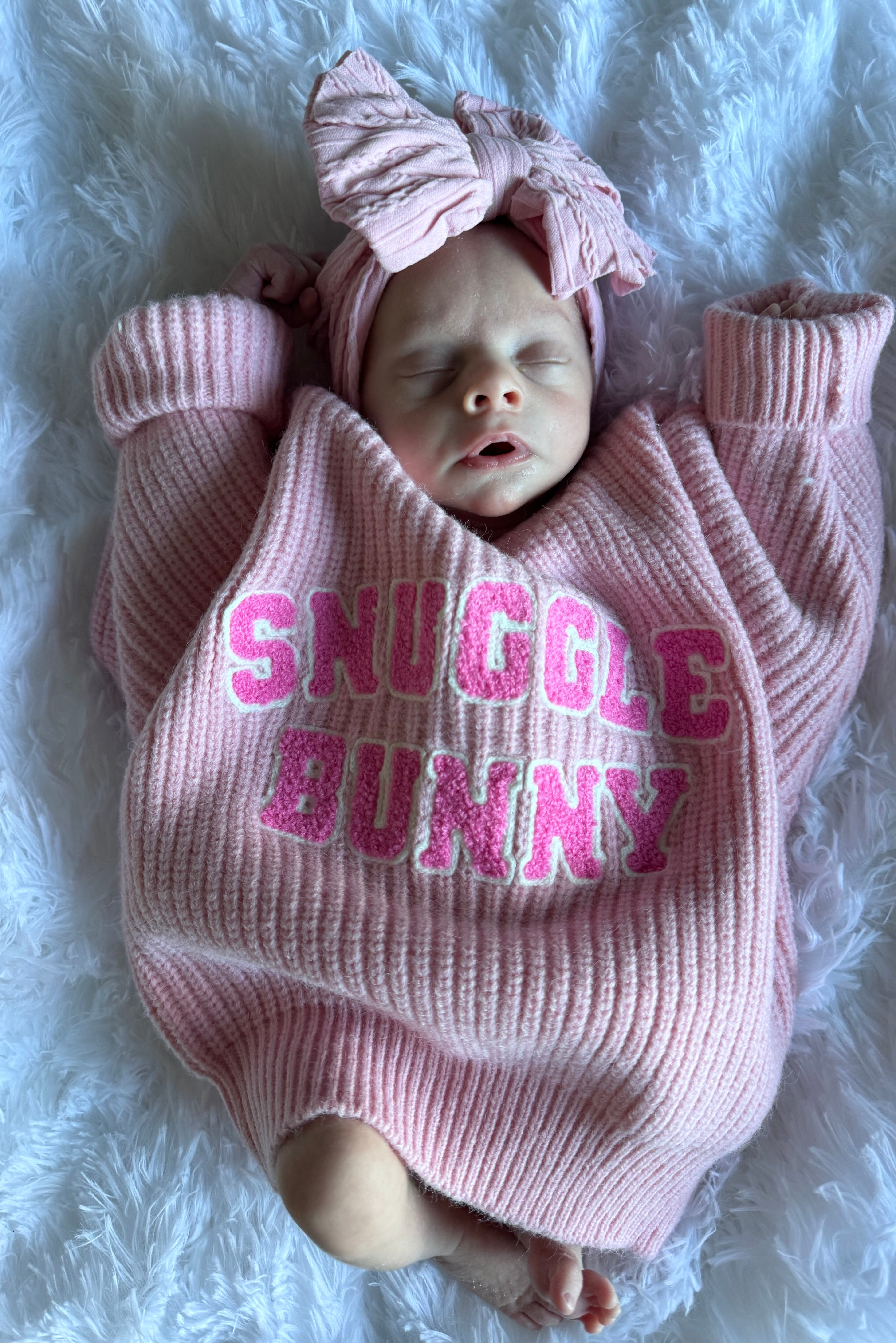 A sleeping baby in a pink sweater with "Snuggle Bunny" text, wearing a matching headband, on a fluffy white blanket.