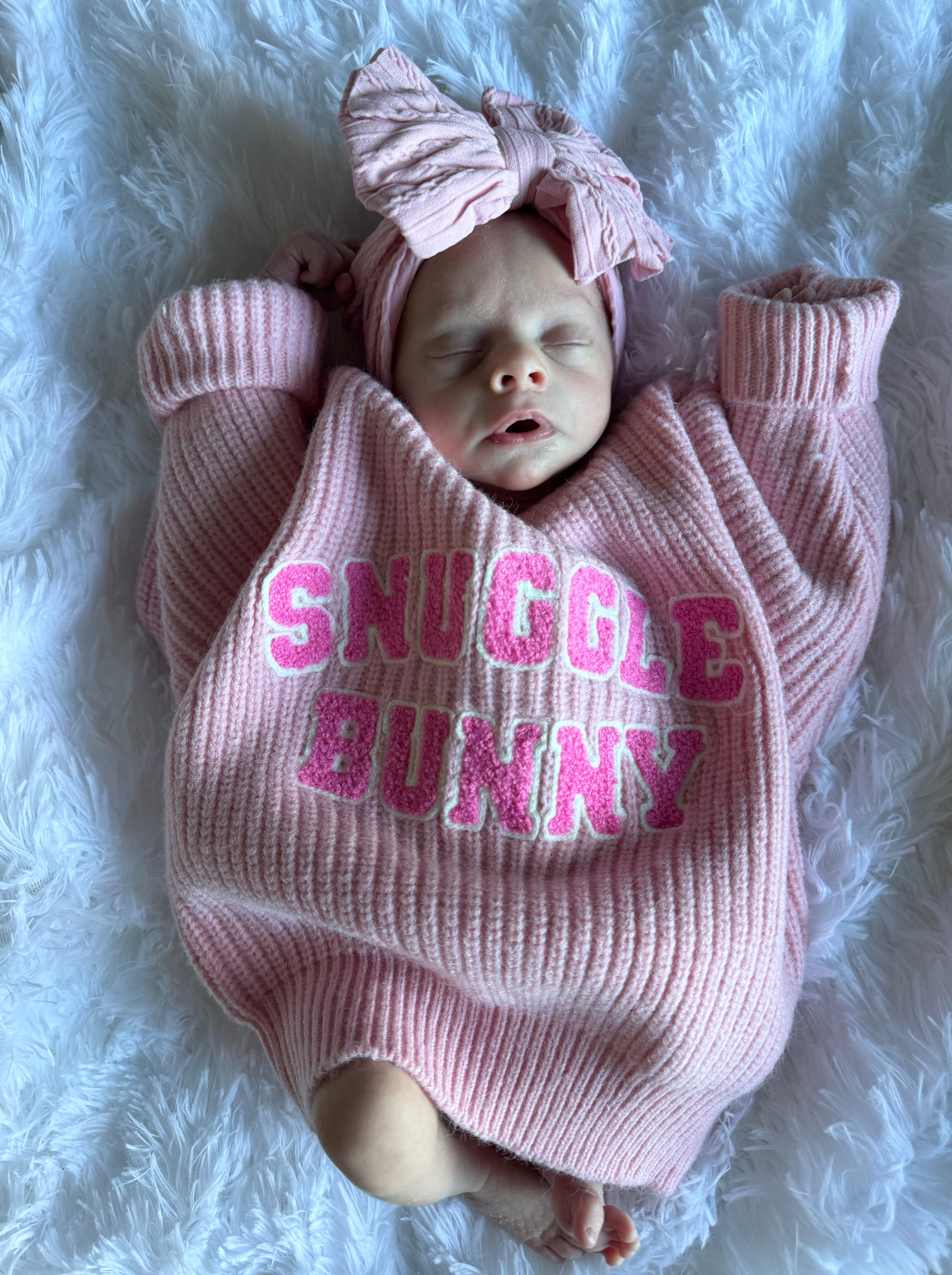 A sleeping baby in a pink sweater with "Snuggle Bunny" text, wearing a matching headband, on a fluffy white blanket.