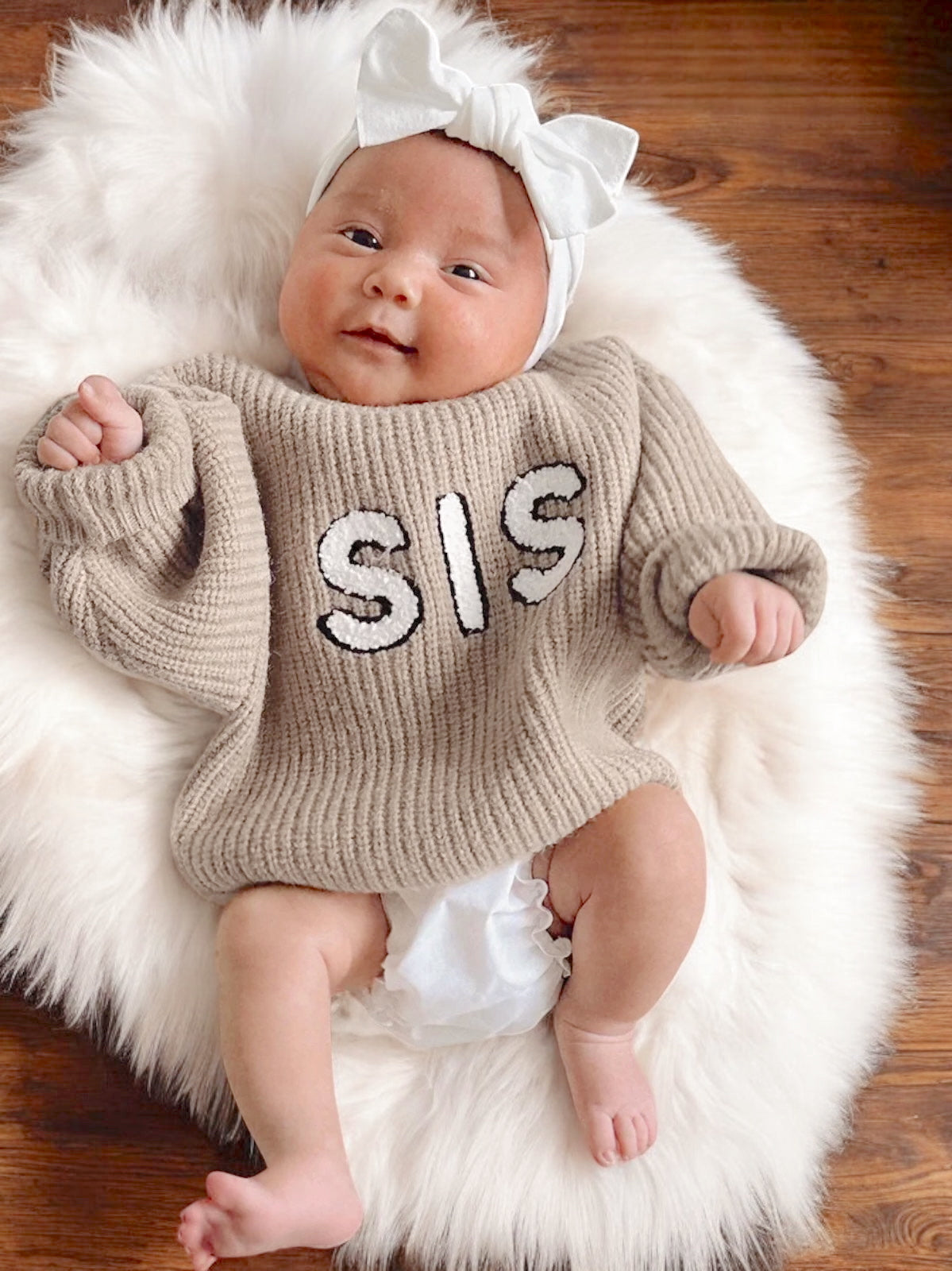 Happy baby wearing a beige sweater with "SIS" text, white bow, lying on a fluffy rug.