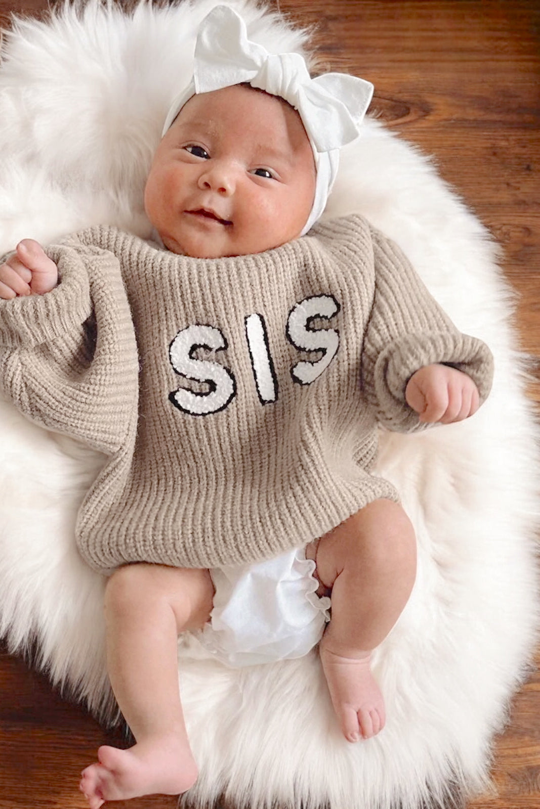 Happy baby wearing a beige sweater with "SIS" text, white bow, lying on a fluffy rug.