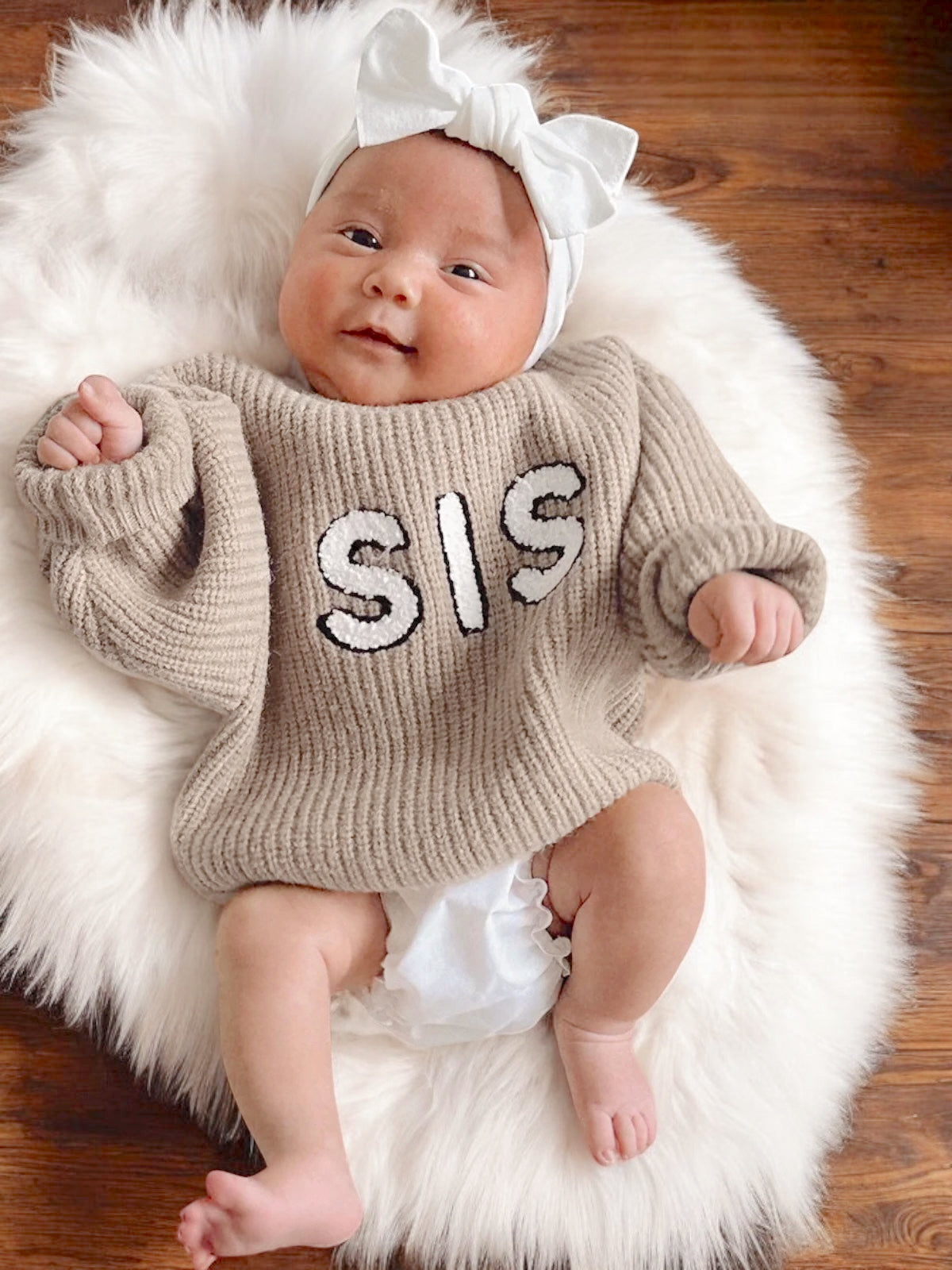Happy baby wearing a beige sweater with "SIS" text, white bow, lying on a fluffy rug.
