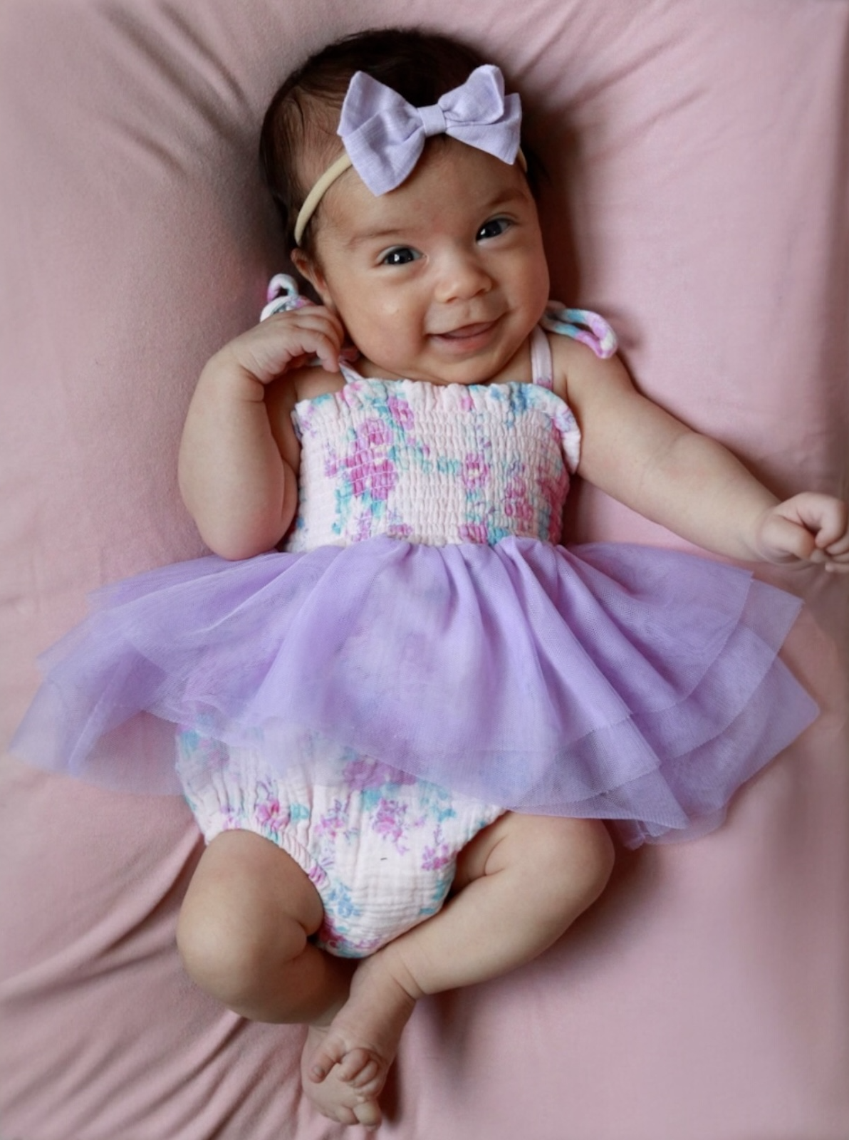 Smiling baby girl in a floral dress with a purple tutu and headband, lying on a pink background.