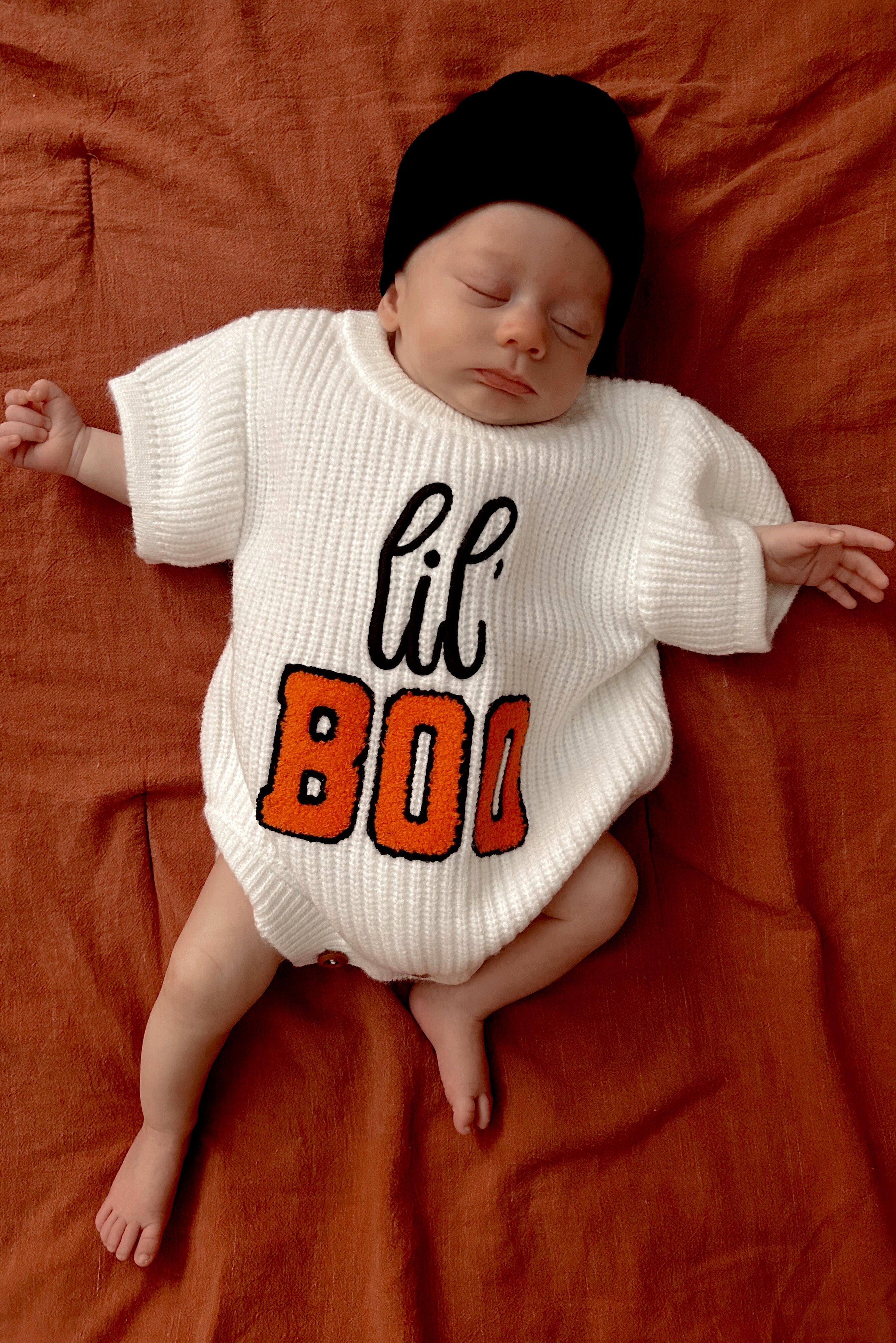Infant wearing a white sweater with "lil' BOO" in orange, sleeping on an orange fabric background.