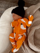 Baby in an orange ghost-patterned onesie sleeps on a soft blanket, wearing a black pom-pom beanie.