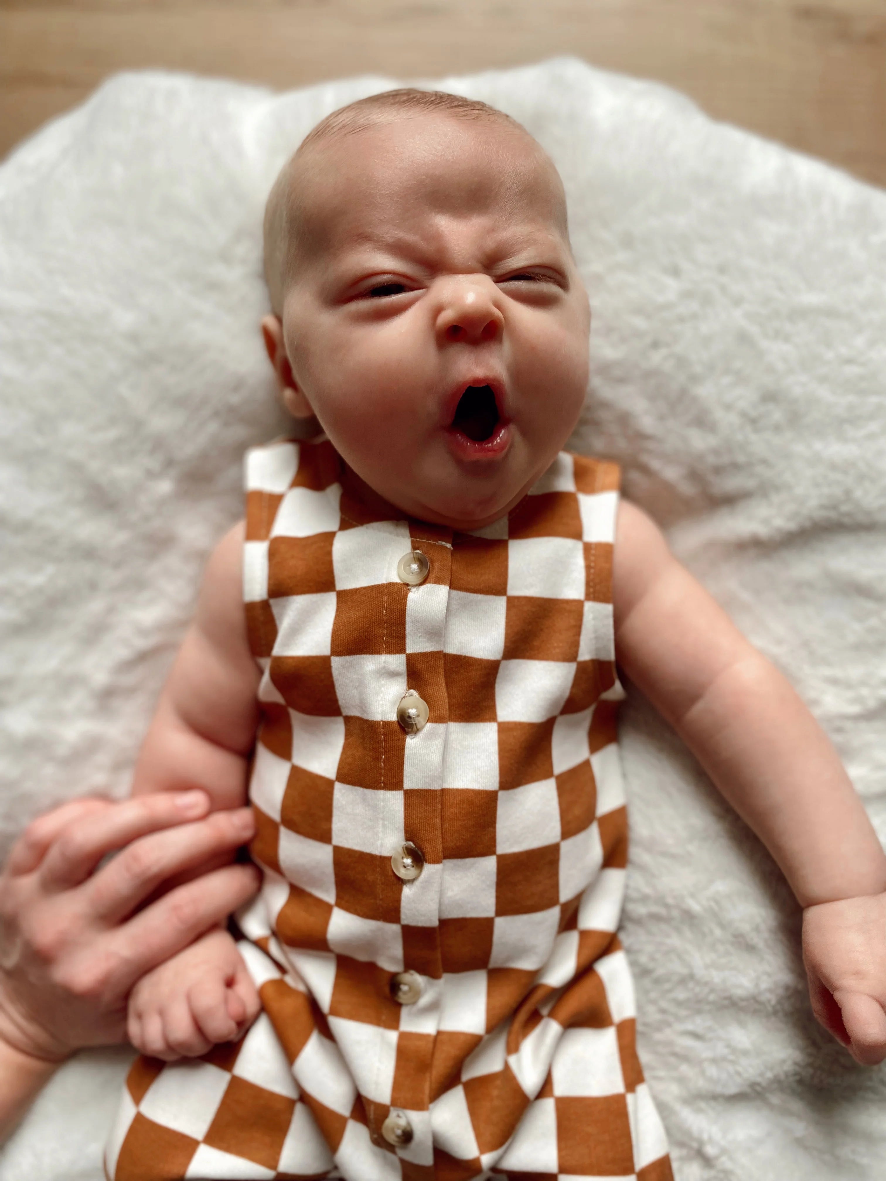Baby dressed in a brown and white checkered outfit yawning on a soft blanket, with a hand gently holding their arm.
